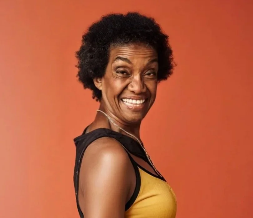 Smiling woman with short curly hair, wearing a yellow and black top, against an orange background.