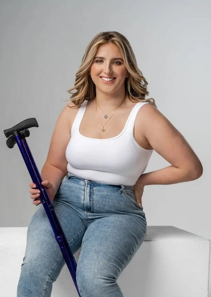 A young woman with shoulder-length blonde hair, wearing a white tank top and blue jeans, sitting on a white surface, holding a blue cane, smiling at the camera.