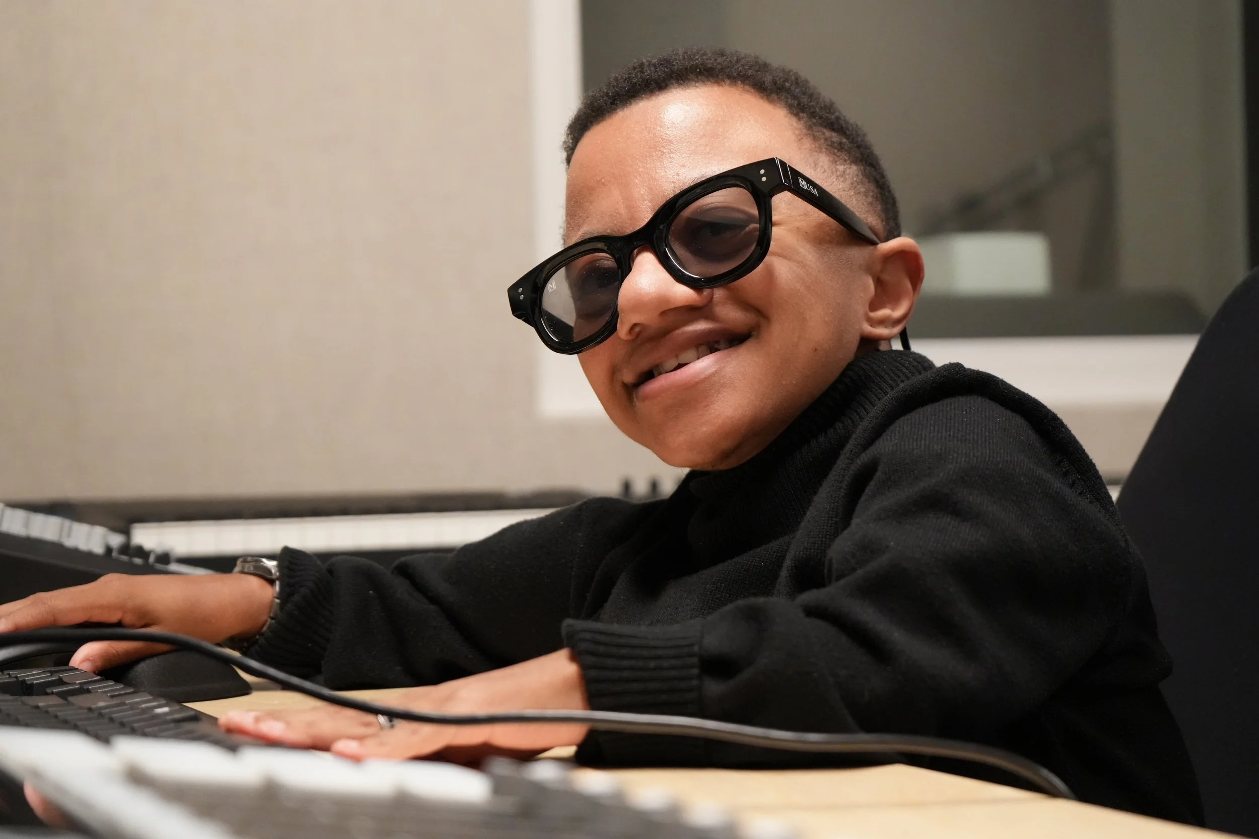 A young man of with short curly hair wearing black sunglasses and a black sweater, smiling while playing a keyboard in an indoor setting.