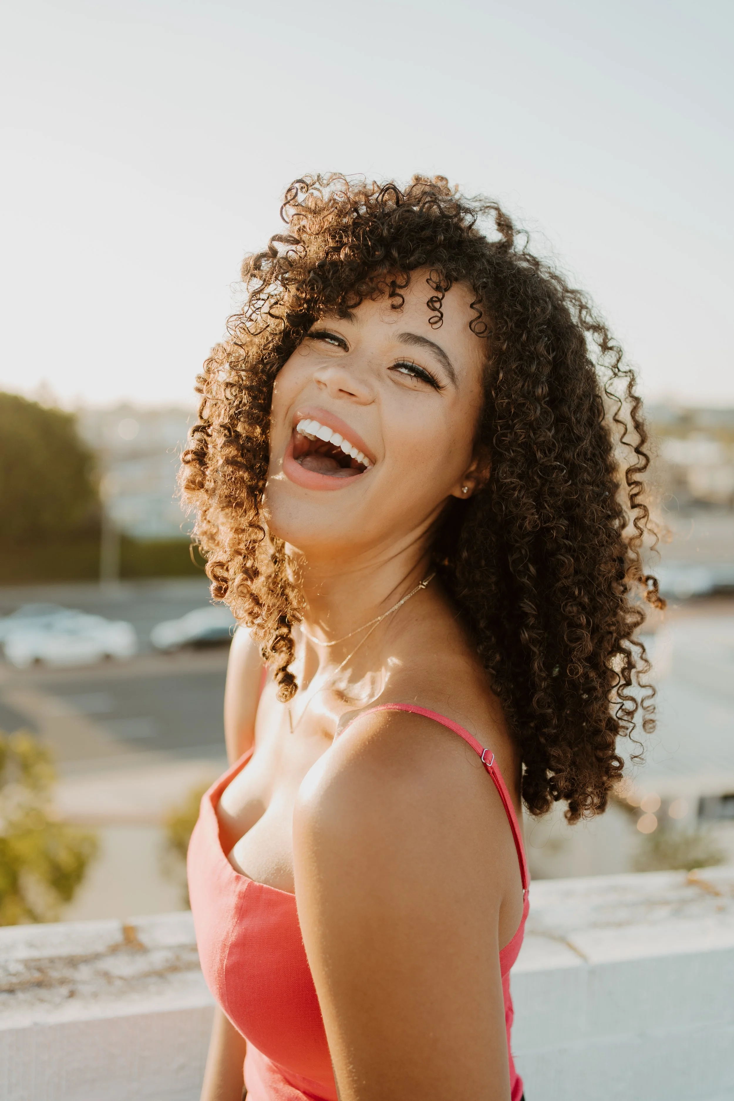 A woman with curly hair and a bright smile, wearing a pink spaghetti strap top, standing outdoors during daylight, with blurred cars and trees in the background.
