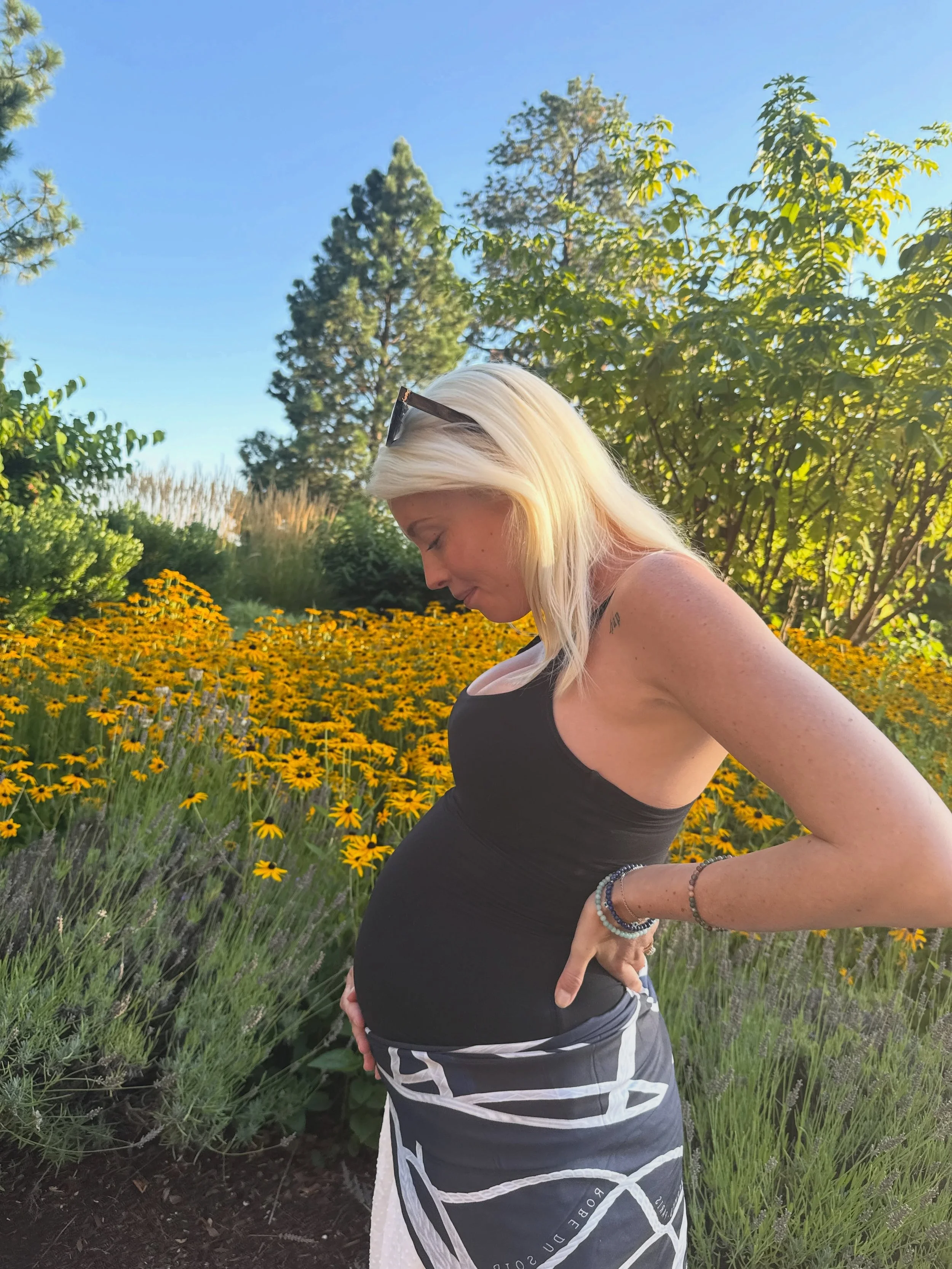 A pregnant woman with blonde hair wearing sunglasses on her head, a black tank top, patterned shorts, and bracelets, standing outdoors in a garden filled with yellow flowers and green trees, during daytime.