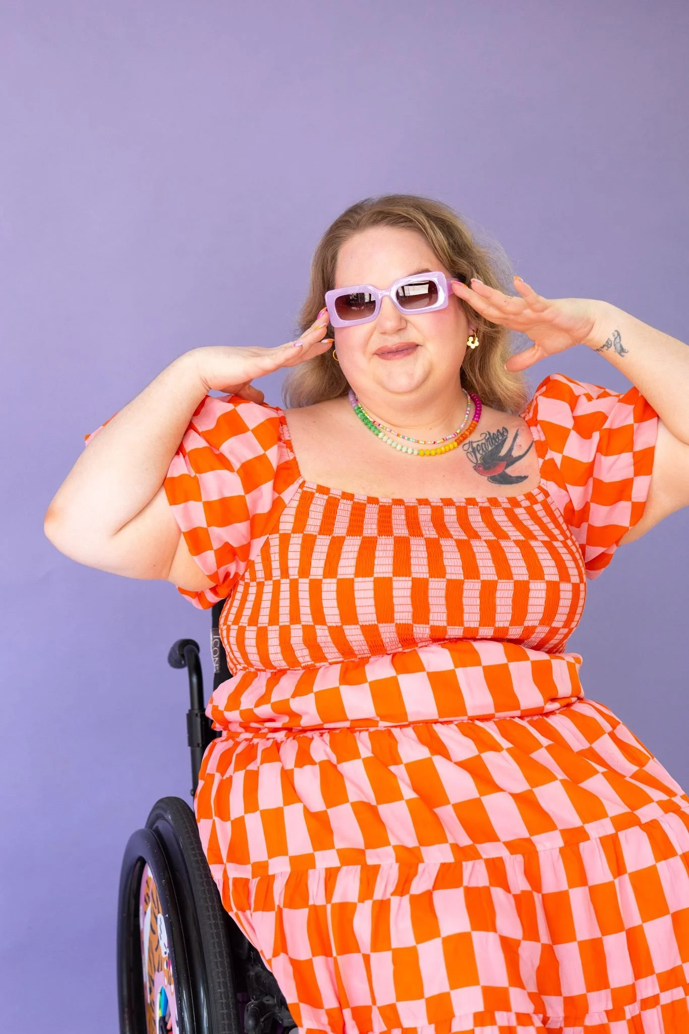 A woman in a wheelchair wearing a bright orange and pink checkered dress, sunglasses, and colorful jewelry, posing with her hands near her face against a purple background.