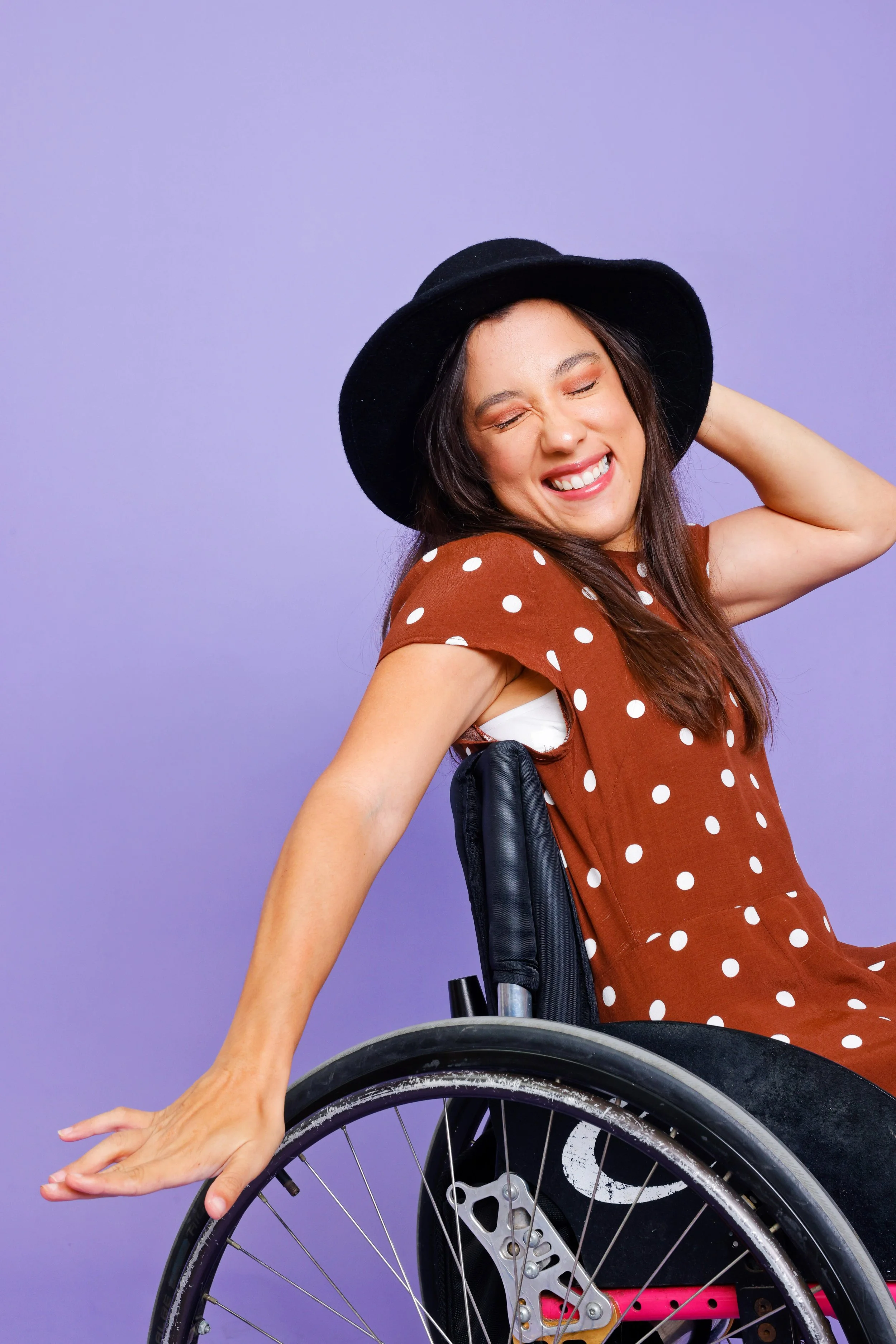 A smiling woman with long brown hair, wearing a black hat and a polka dot brown dress, sitting in a wheelchair, with her eyes closed and one hand resting on the wheel, against a purple background.
