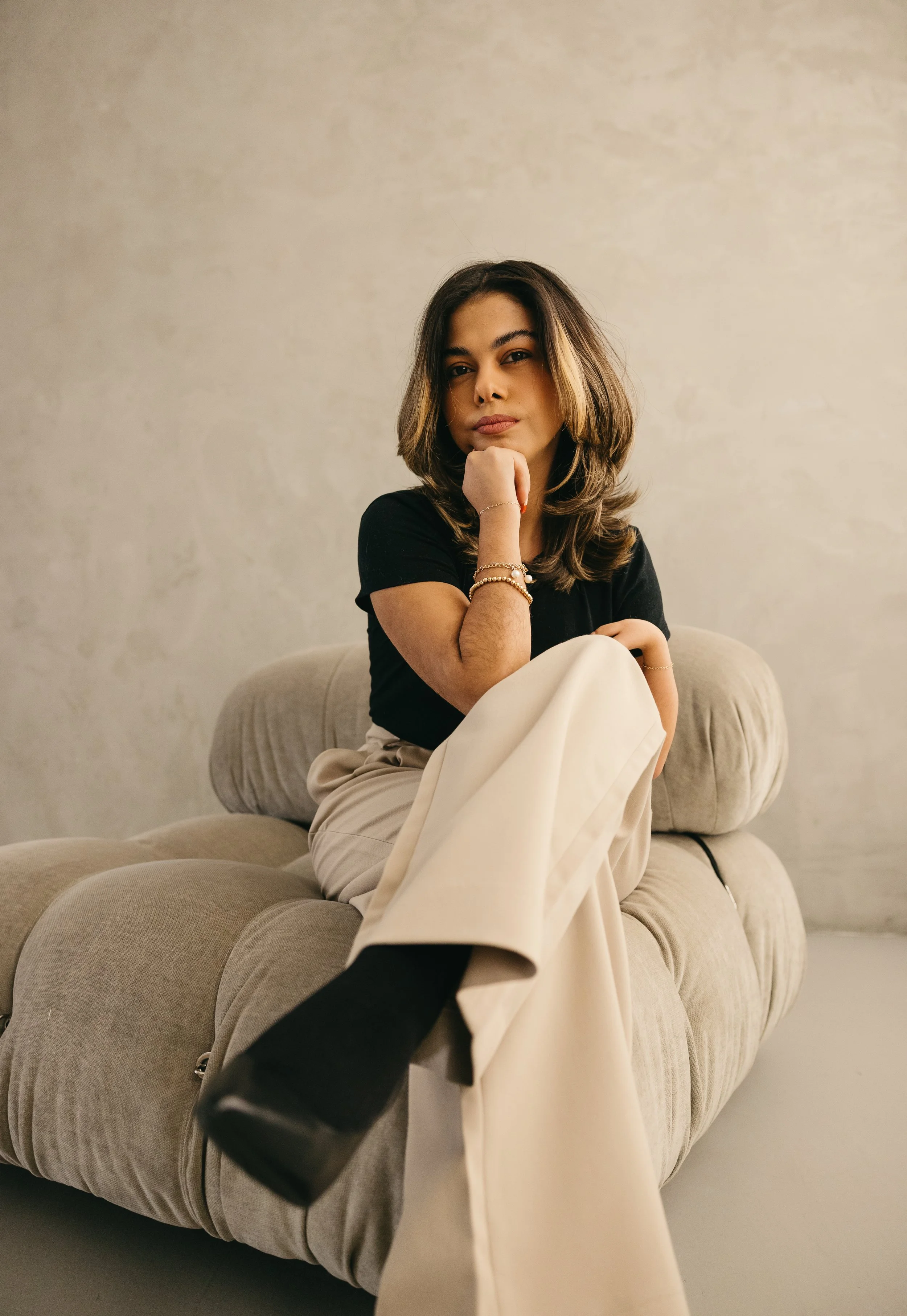 A woman sitting on a beige couch with her legs crossed, wearing a black top, beige pants, black shoes, gold jewelry, and resting her chin on her hand, in a room with a neutral-colored wall.