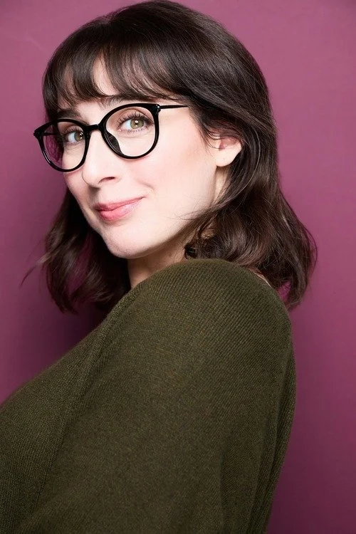A young woman with brown hair, glasses, and a gentle smile, wearing a dark green top, standing in front of a pink background.