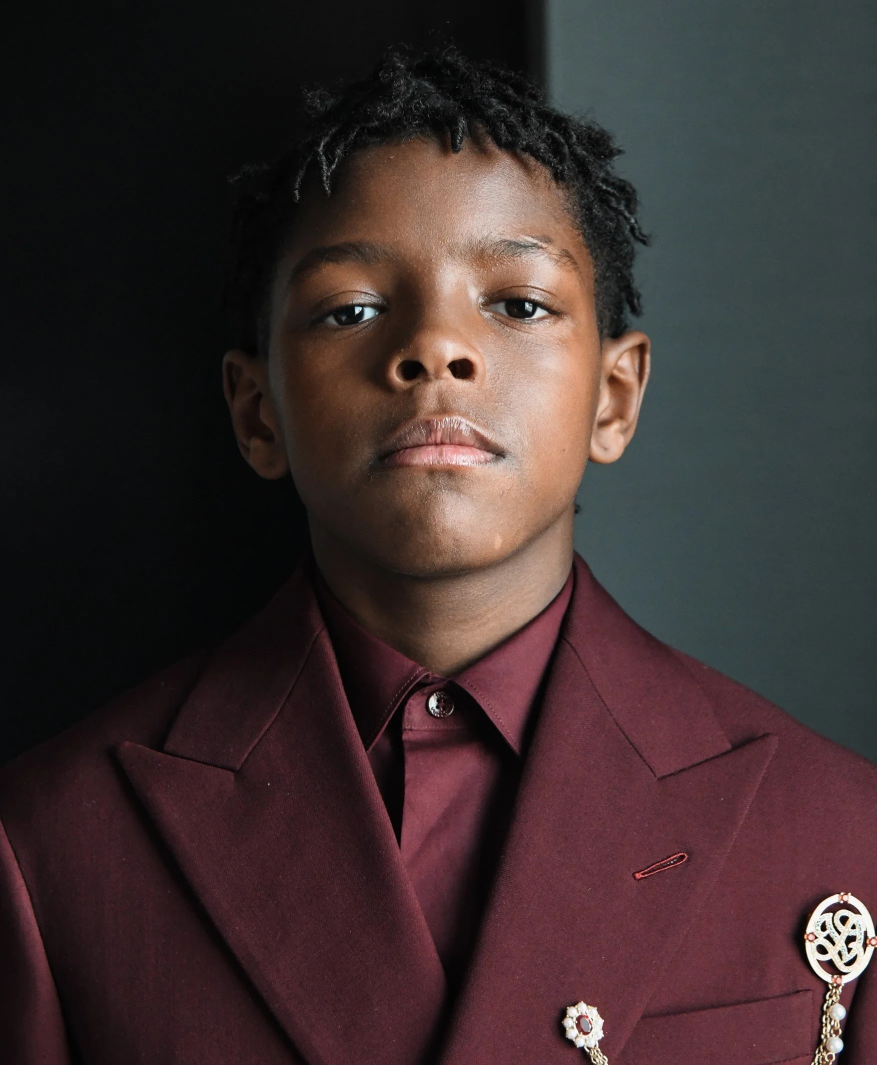 Close-up of a young man with dark skin and short curly hair, wearing a burgundy blazer with decorative pins, against a plain dark background.