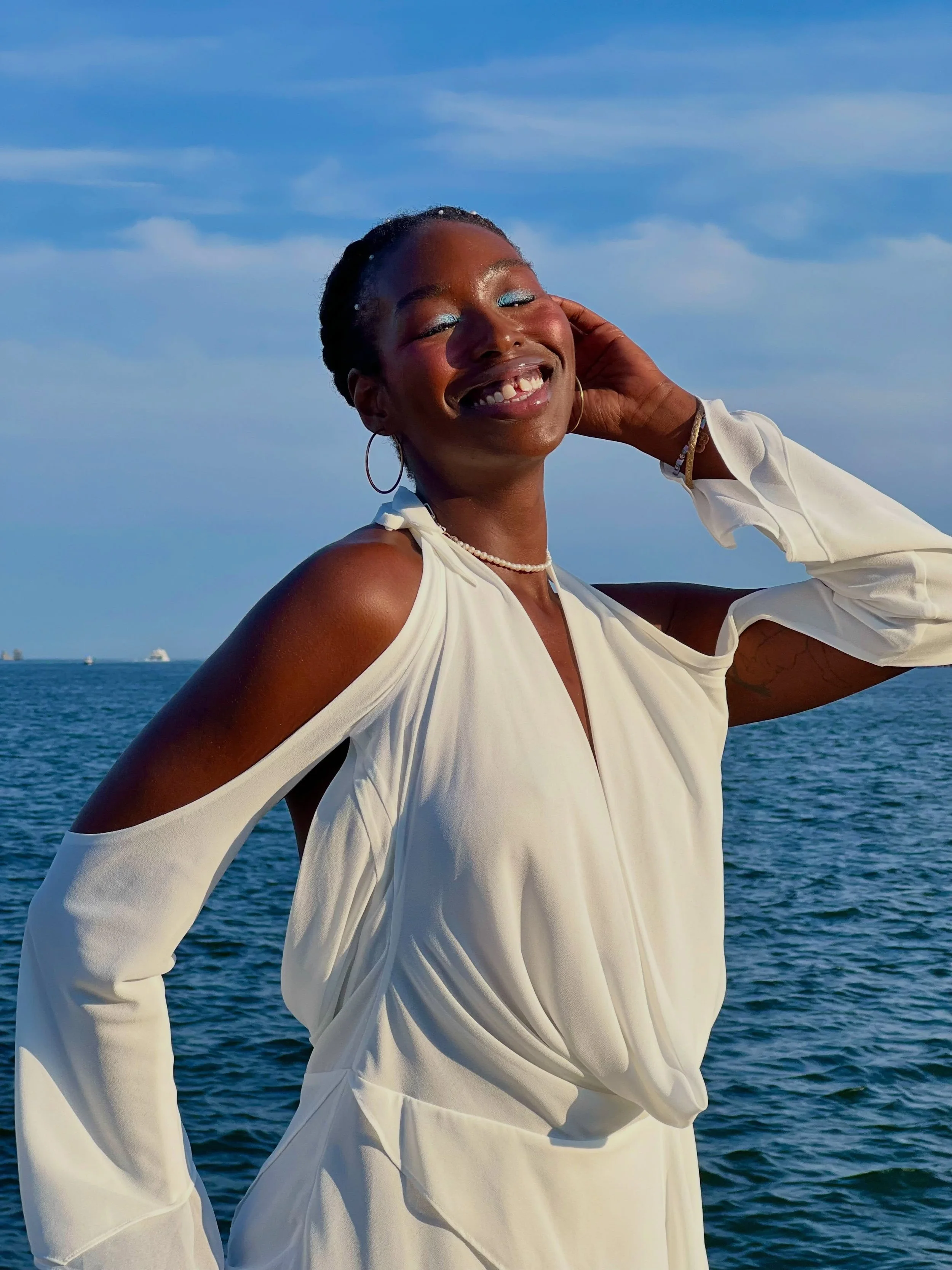 A woman with dark skin, wearing a white dress with cutout shoulders, smiling and posing outdoors near a body of water with ships in the background, under a blue sky with clouds.