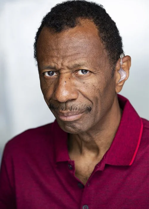 Close-up of an older man with dark skin, short curly black hair, and a mustache, wearing a maroon collared shirt, looking directly at the camera with a serious expression.