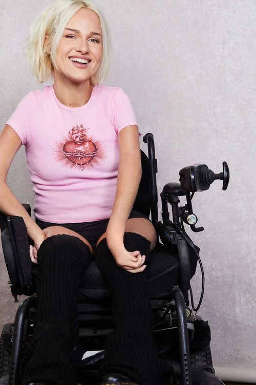 A young woman with blond hair smiling, sitting in a motorized wheelchair, wearing a pink t-shirt with a heart graphic, black striped thigh-high socks, and black shorts.