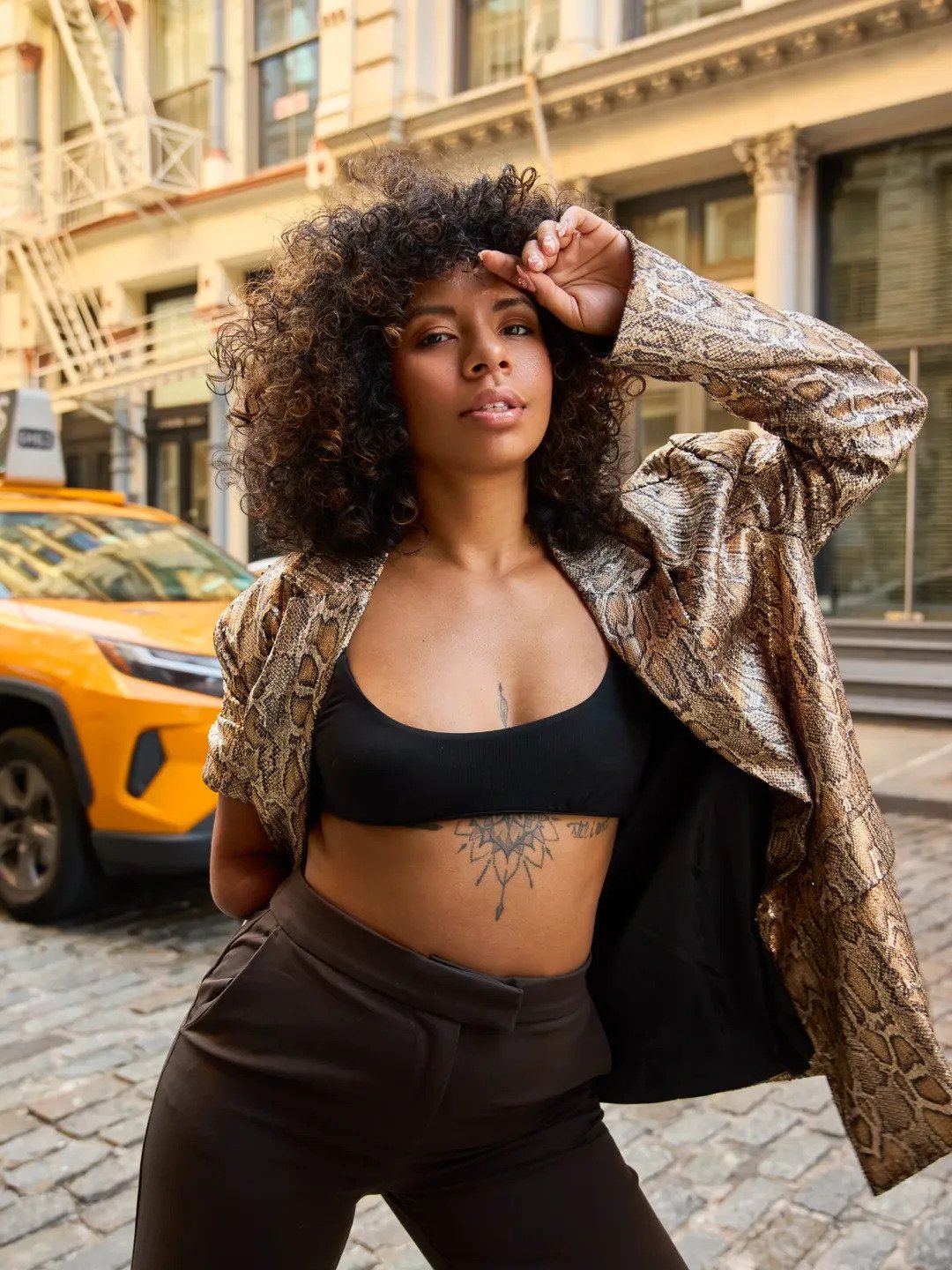 A woman with curly hair wearing a black crop top and netted black pants, standing on a cobblestone street in front of a yellow taxi. She has a snake-skin patterned jacket draped over her shoulders and tattoos on her chest and stomach. She is posing with her right hand raised to her forehead, looking directly at the camera.