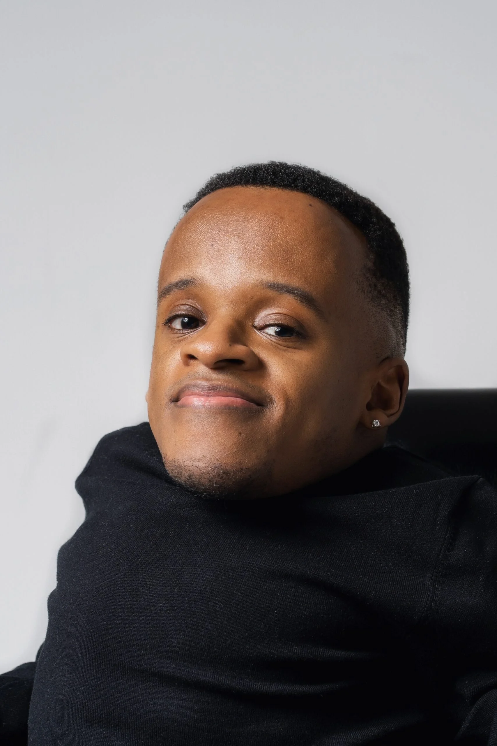 Close-up of a person with short hair, a diamond earring, and wearing a black hoodie, smiling slightly. The person has light brown skin and is set against a plain, light-colored background.