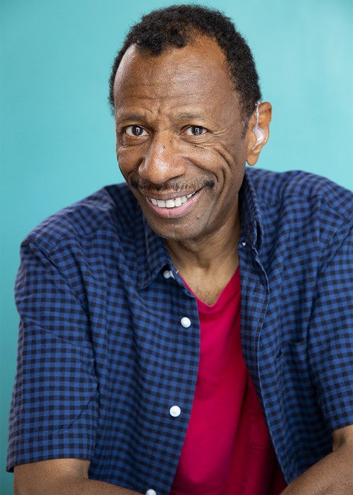 A middle-aged man with short curly hair and brown skin, smiling and wearing a blue checkered shirt and a red T-shirt, against a turquoise background.