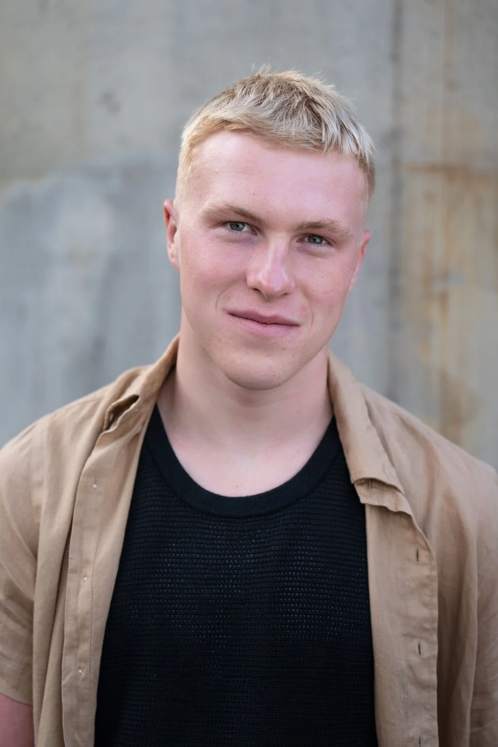 A young man with blond hair and blue eyes looking at the camera with a slight smile, standing outdoors against a grey wooden background.