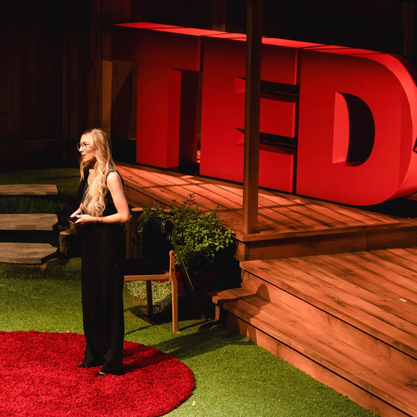 A woman with long blonde hair, wearing glasses and a black dress, is standing on a red circular rug at a TED Talk event. She is smiling and holding a remote, with a TED logo in large red letters on a wooden stage behind her.
