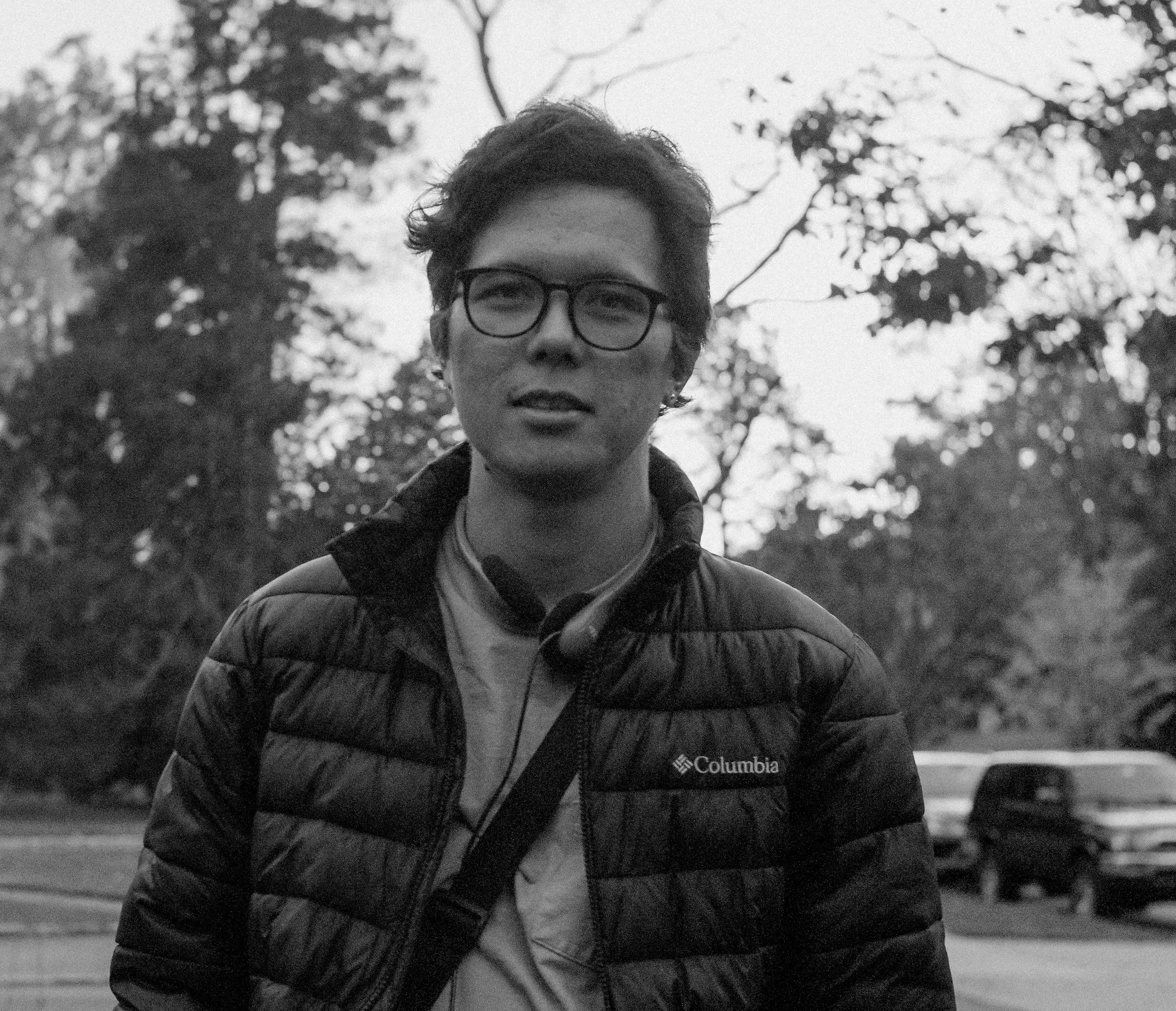 A young man with glasses, curly hair, and a Columbia jacket stands outdoors during twilight or early evening. Trees and a few birds in flight are visible in the background.
