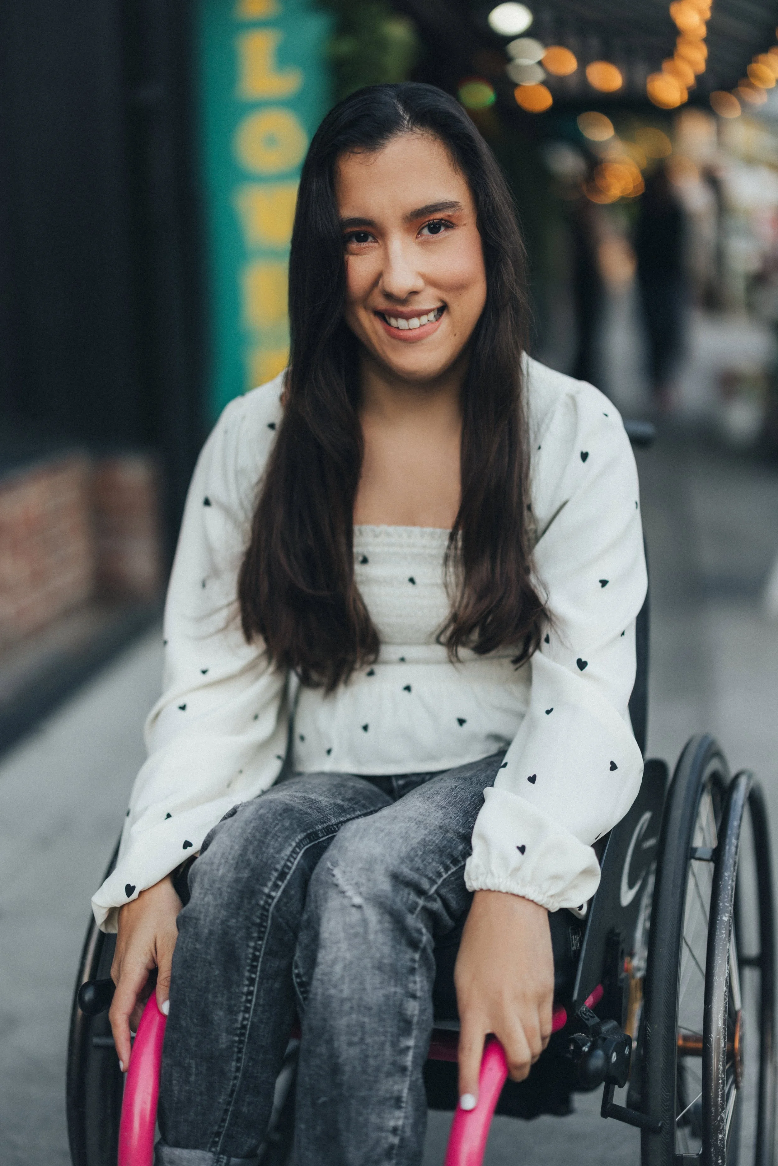 A young woman smiling while sitting in a wheelchair on a city street at dusk, holding pink handrails, with blurred city lights in the background.