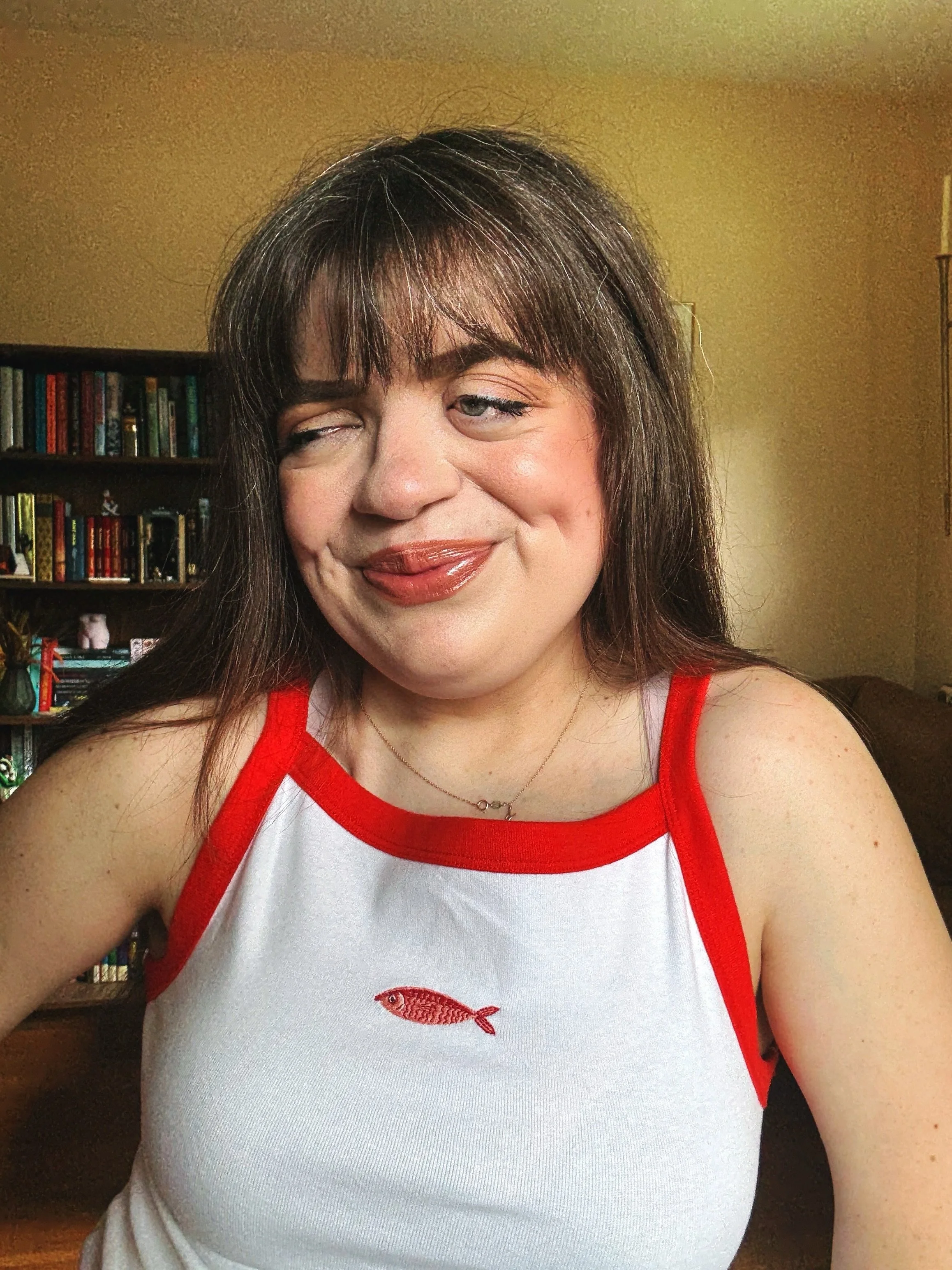 Close-up of a woman with long brown hair wearing a white tank top with red trim and a small red fish embroidery. She is smiling and winking. In the background, there is a bookshelf filled with books and a beige wall.