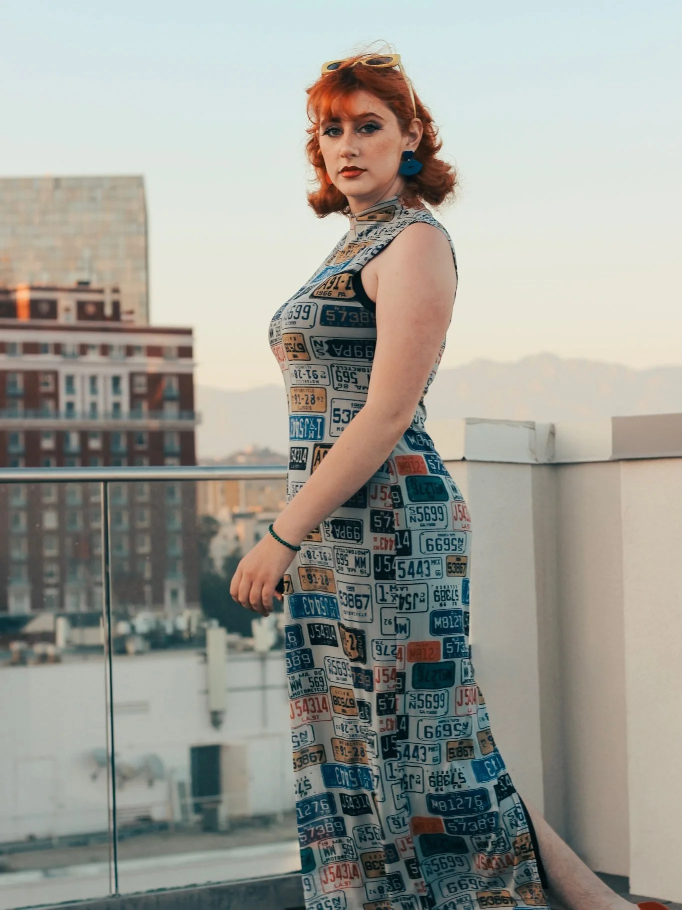 A woman with red hair wearing a dress made of various colorful license plates stands on a rooftop with city buildings in the background.