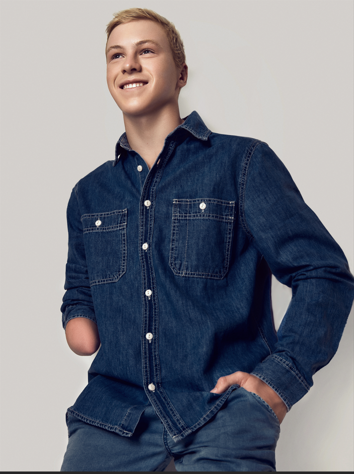 young man smiling, wearing a denim shirt and jeans, standing with one hand in pocket, against a plain background