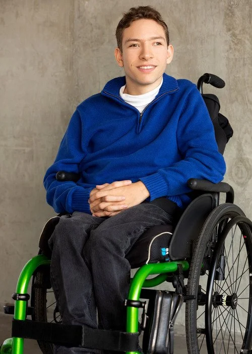 A young man with short brown hair sitting in a wheelchair against a plain gray wall, wearing a blue zip-up hoodie and smiling.