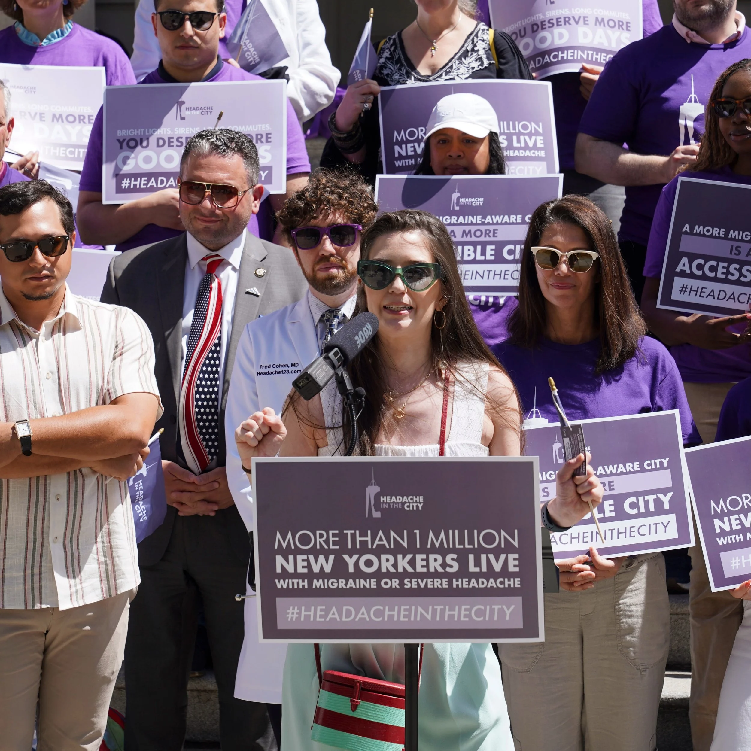 A group of people participating in a protest or rally holding signs about migraine awareness and headache relief in an outdoor setting. Some signs read 'More than 1 million New Yorkers live with migraine or severe headache' and 'You deserve good days