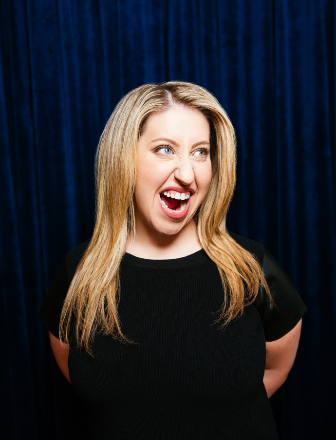 A woman with blonde hair wearing a black top, making a playful facial expression with one eye squinted and mouth open, against a dark blue curtain background.