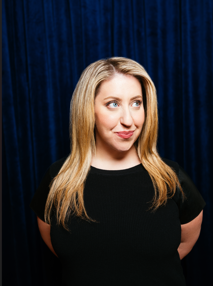 A woman with long blonde hair and blue eyes standing in front of a dark blue curtain, wearing a black top, looking to the side with a slight smile.