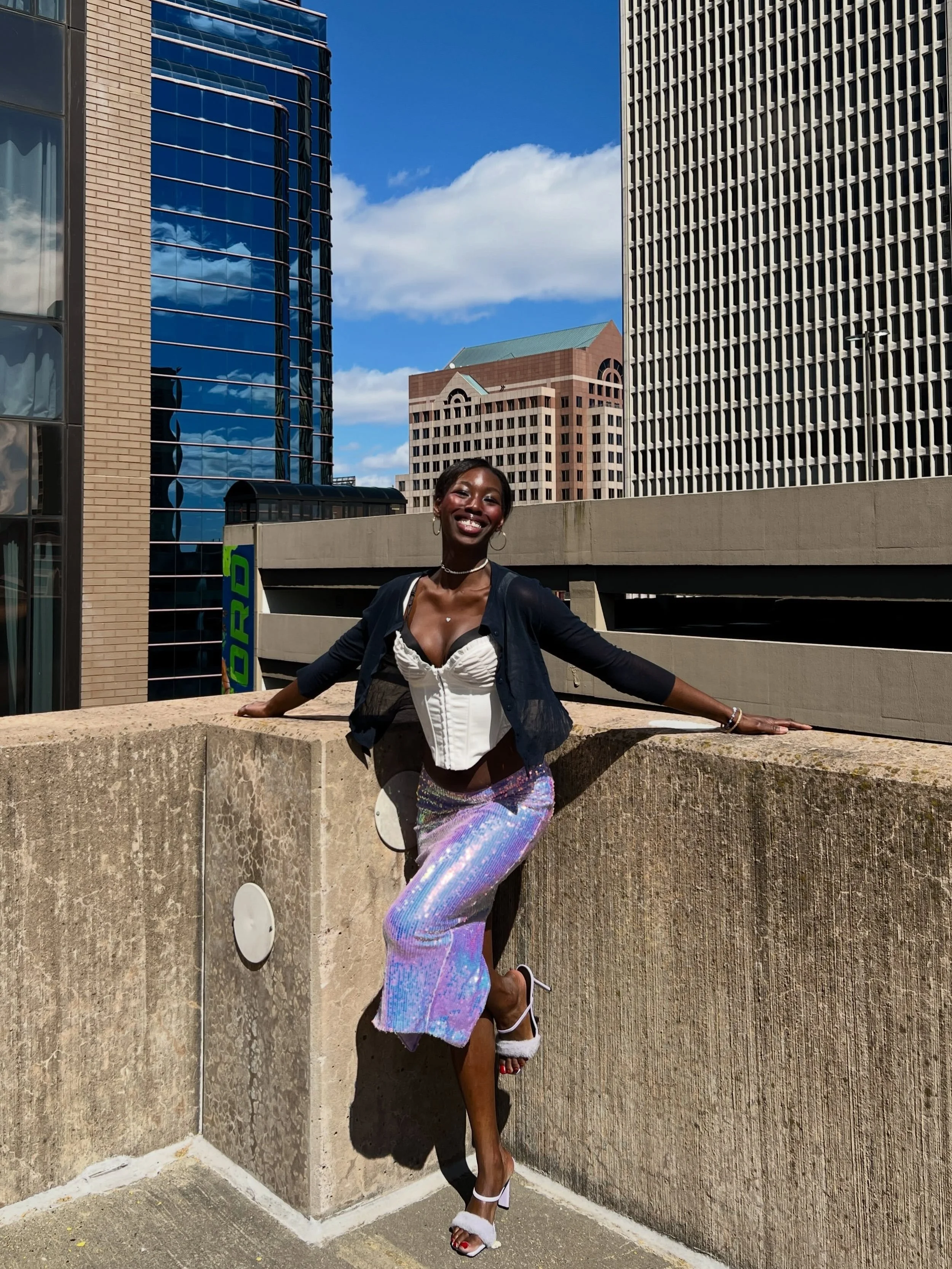 A woman smiling and posing on a city rooftop with tall buildings in the background, wearing a fashionable outfit and high heels.