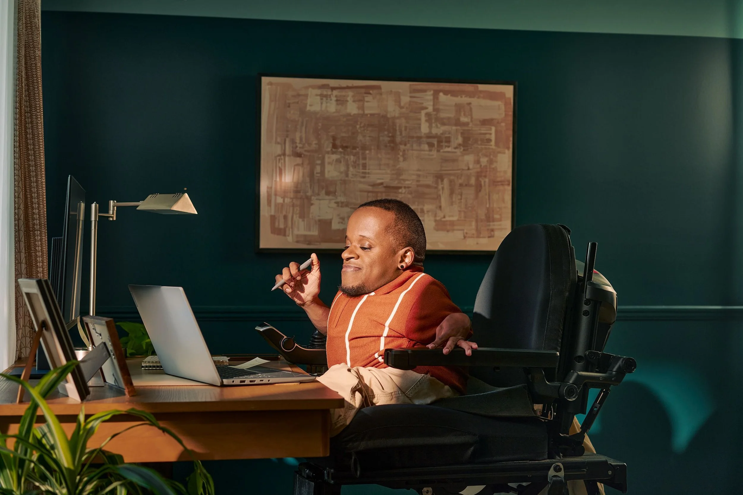 A man in a wheelchair working at a desk in an office, with a green wall and framed artwork behind him, and a lamp and plant visible in the foreground.