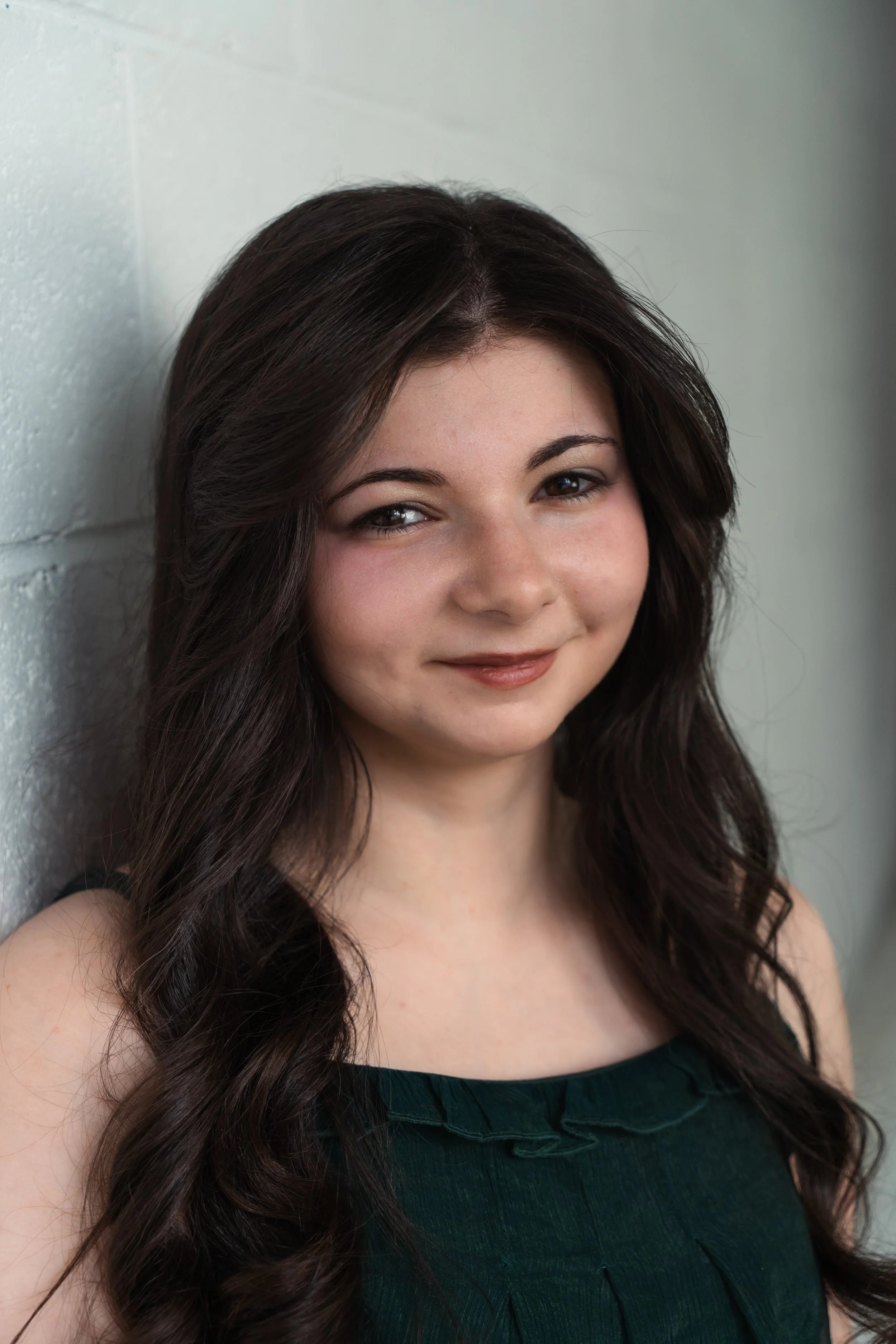 Close-up of a young woman with long, dark, wavy hair, smiling and leaning against a light-colored brick wall, wearing a dark green sleeveless top.