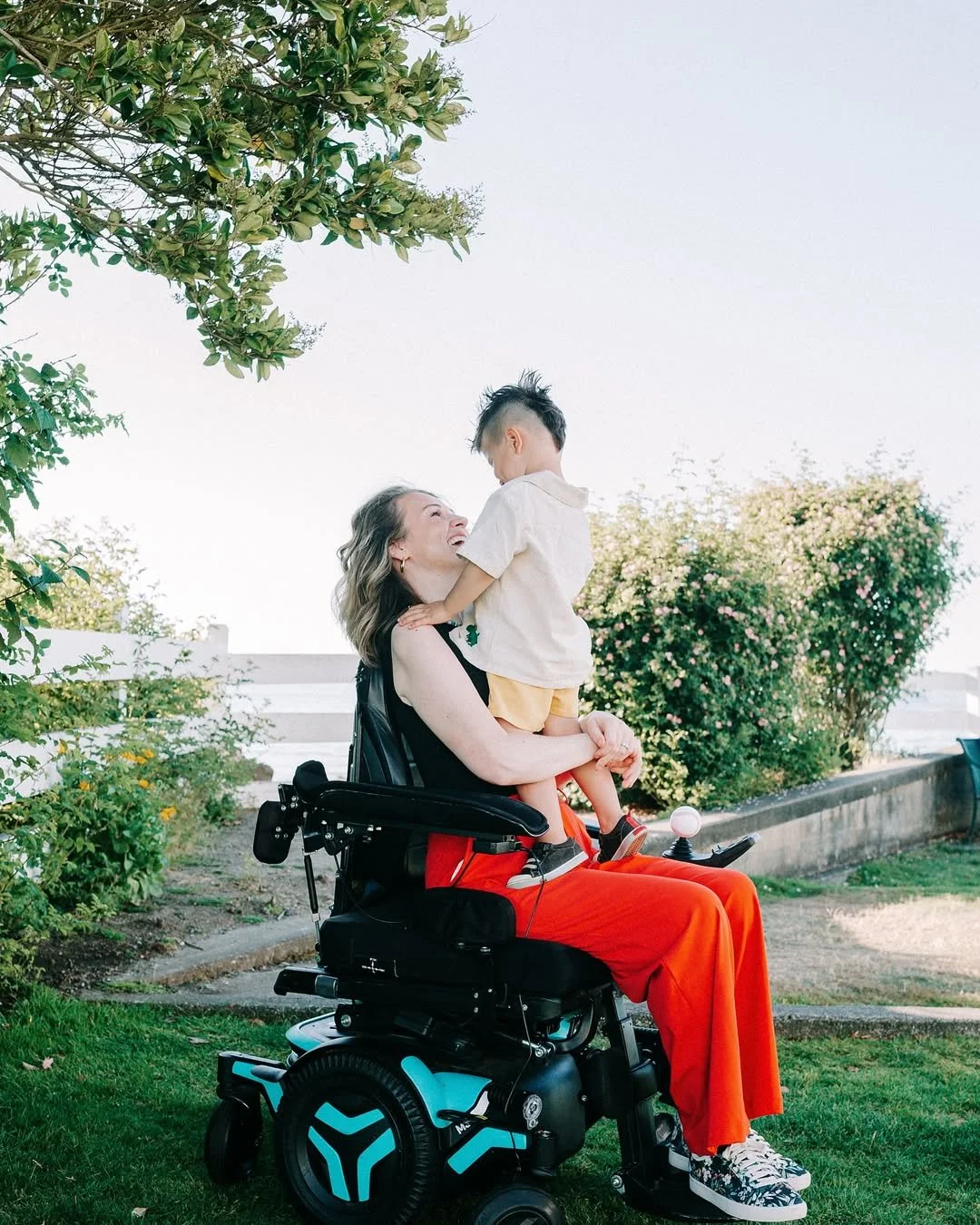 A woman in a motorized wheelchair is holding a young boy on her lap, both smiling and looking at each other, outdoors with trees and bushes in the background.