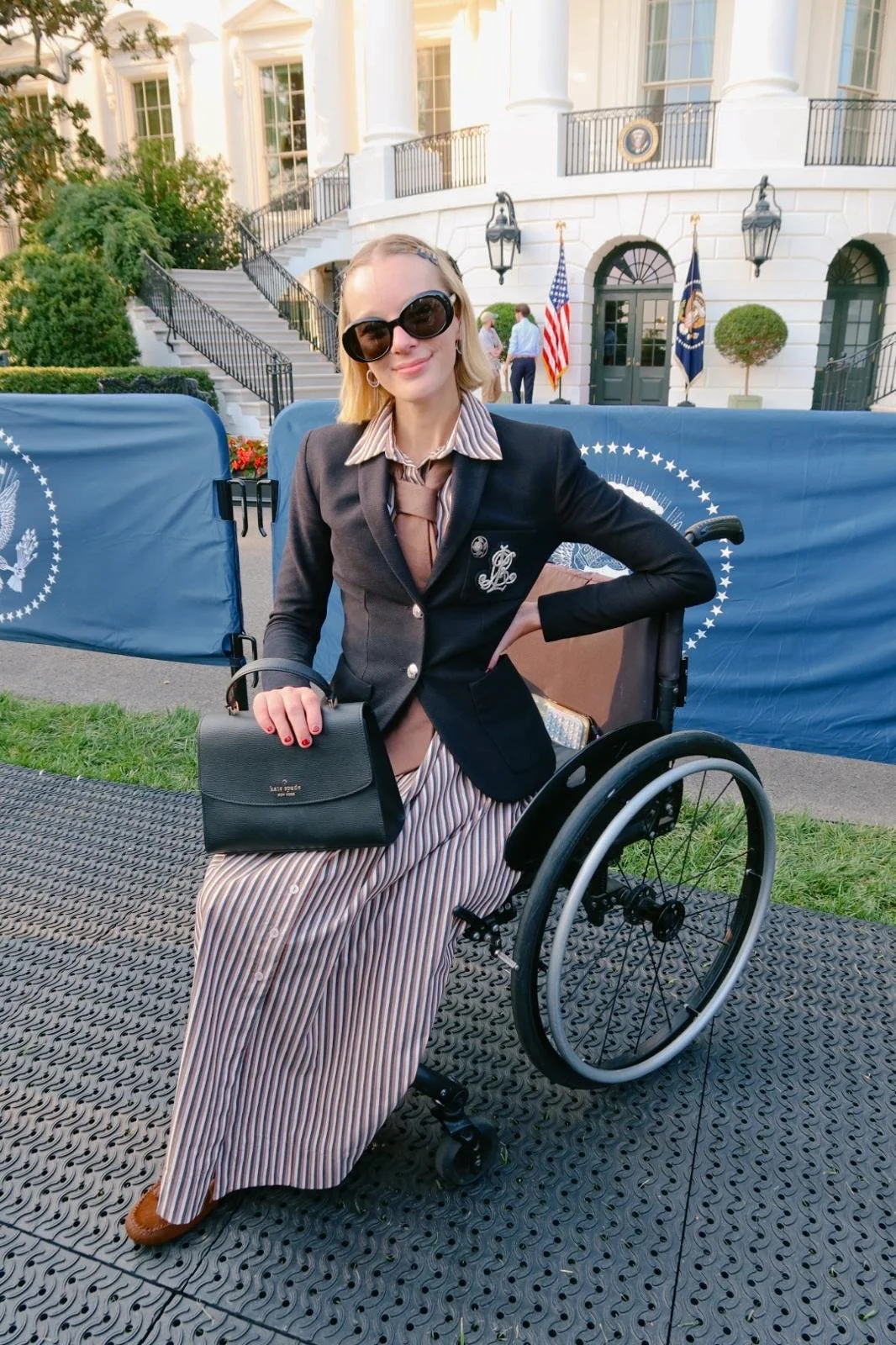 Woman in sunglasses and a suit sitting in a wheelchair outdoors in front of a White House-like building with flags.