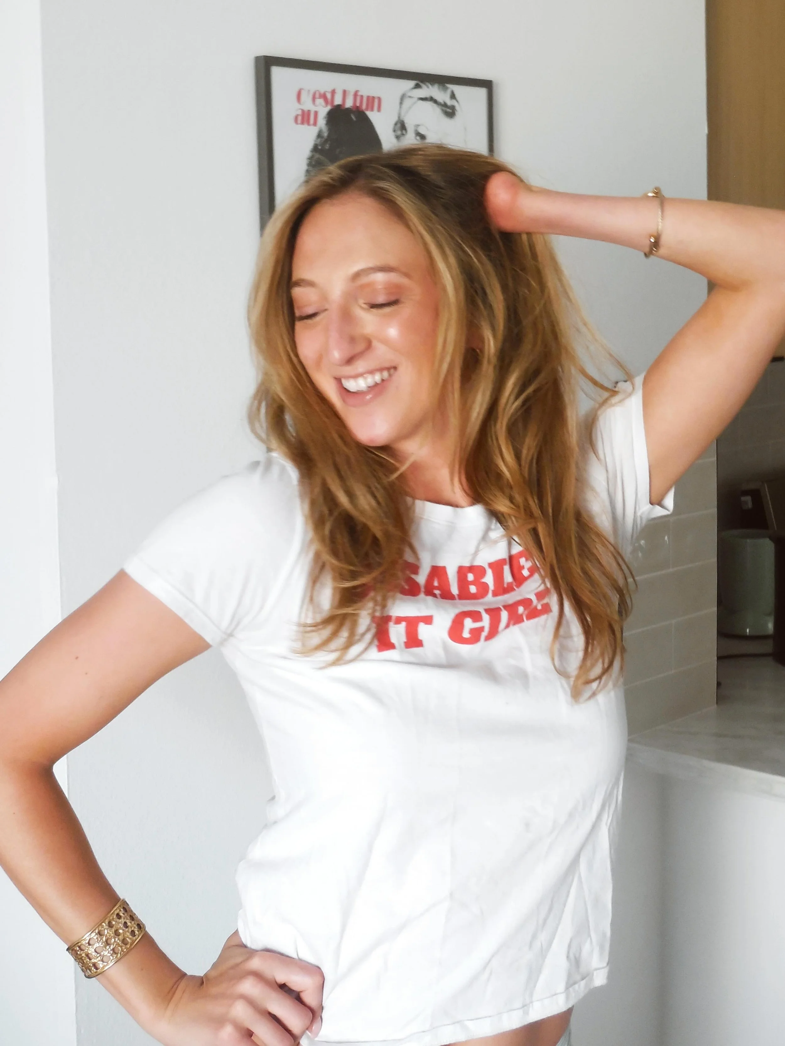 A woman with long blonde hair smiling with closed eyes, wearing a white T-shirt with red text and a gold bracelet, with a limb difference, standing indoors against a white wall with a framed black-and-white illustration in the background.