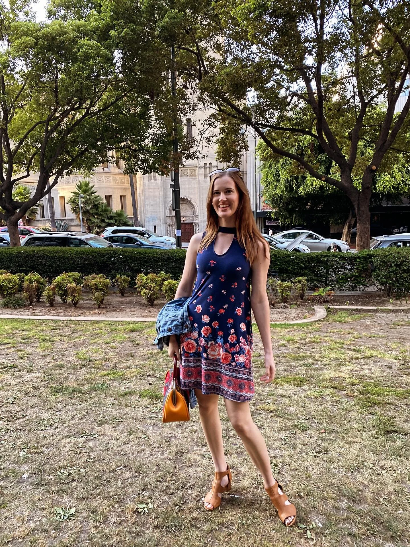 A woman with red hair smiling, wearing a floral dress, tan heels, holding a yellow bag, with a denim jacket over her arm, standing in a park with trees, bushes, and parked cars in the background.