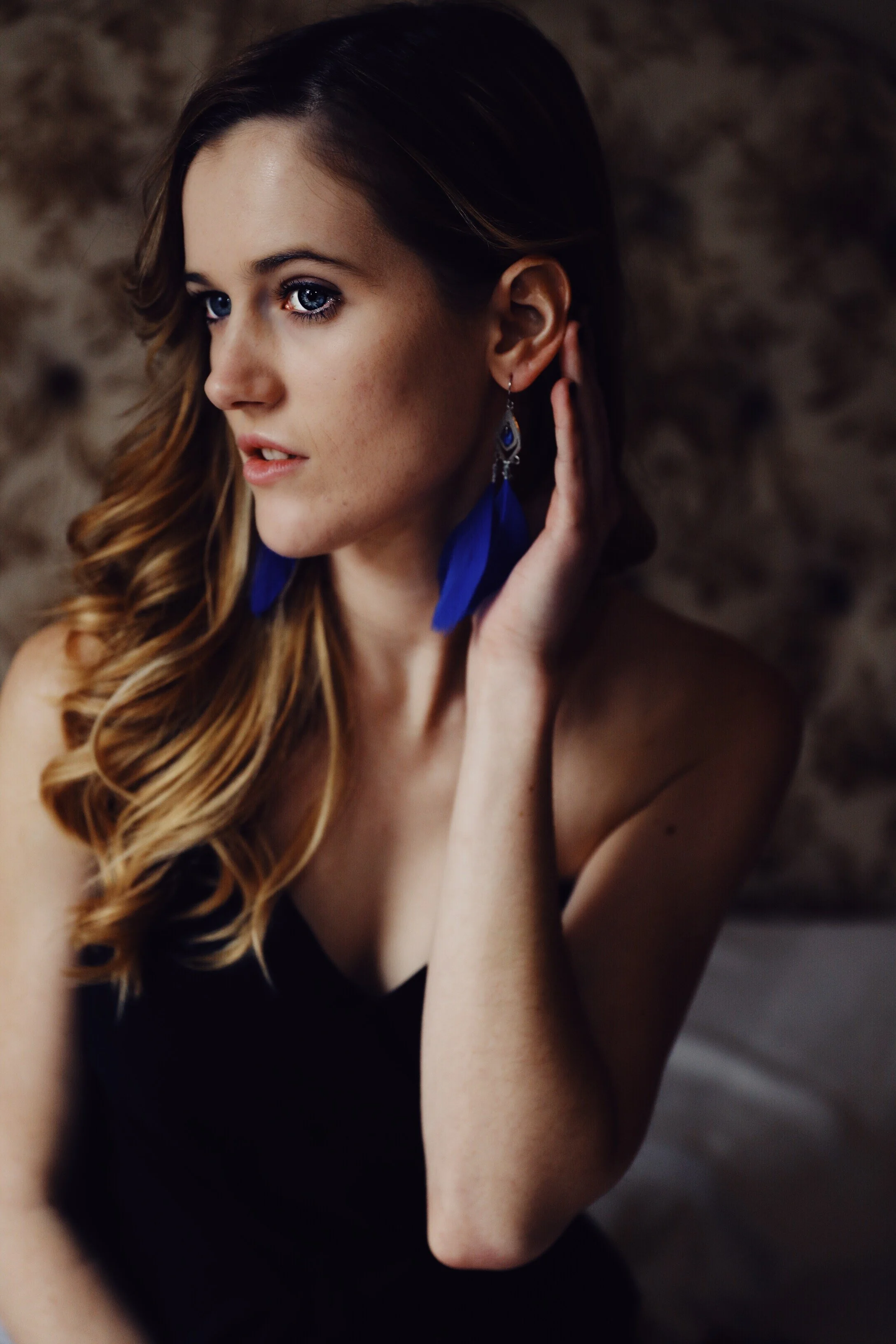 A woman with long, wavy brown hair and blue eyes, wearing peacock feather earrings and a black top, touching her ear with one hand.