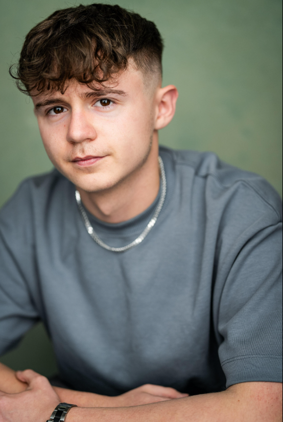 Close-up portrait of a young man with short curly brown hair, wearing a gray t-shirt and a silver chain, against a plain green background.