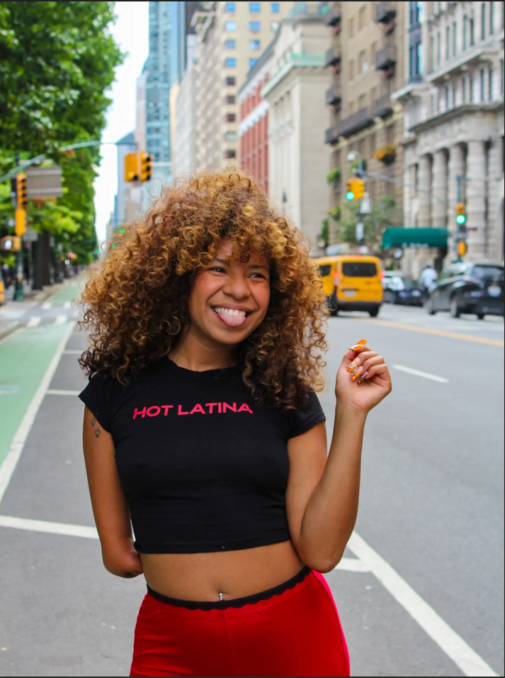 Smiling woman with curly hair standing on city street with buildings, cars, and traffic lights in the background.