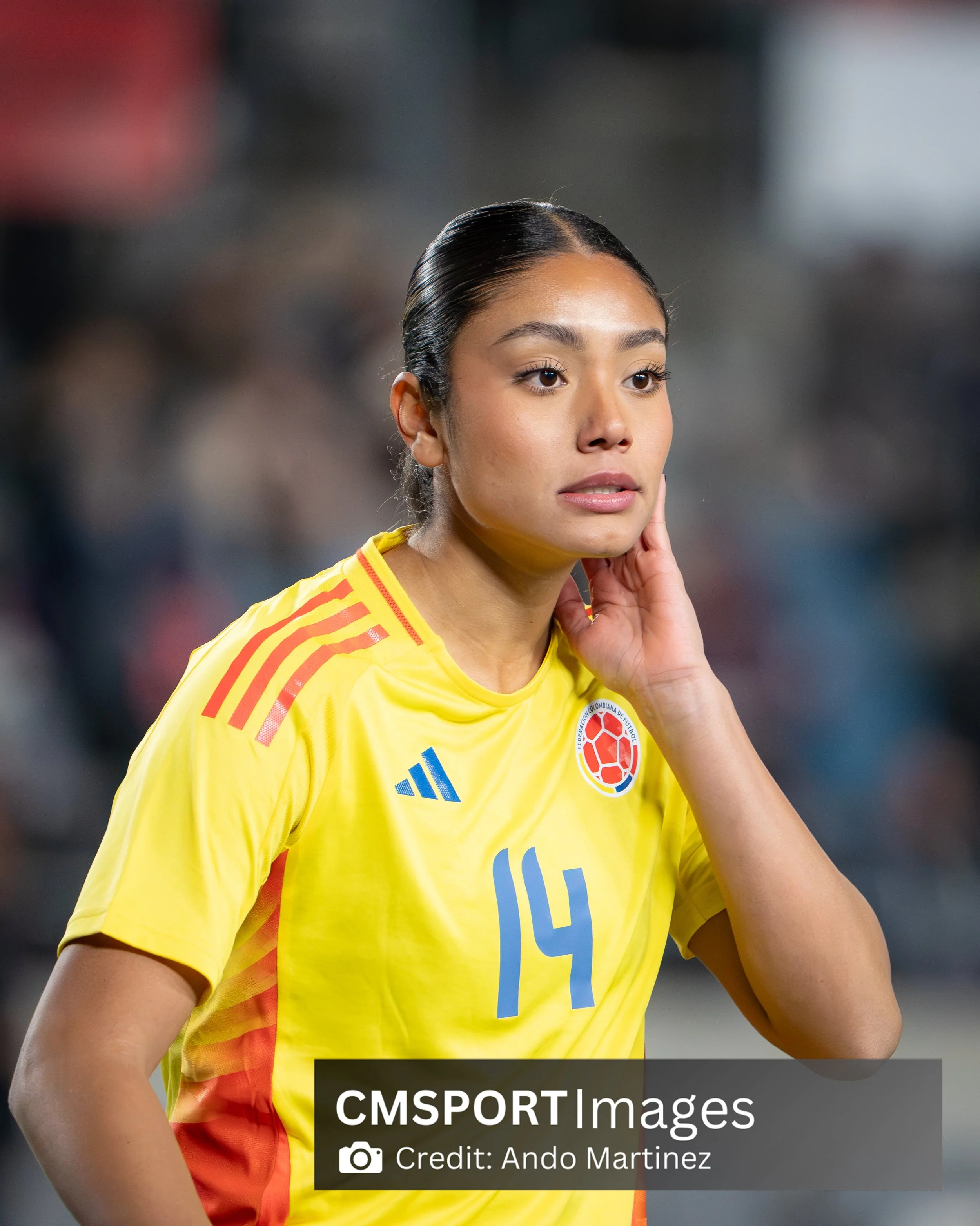 A female soccer player wearing a yellow jersey with the number 14 and the Colombian national team emblem, standing on the field with a focused expression.