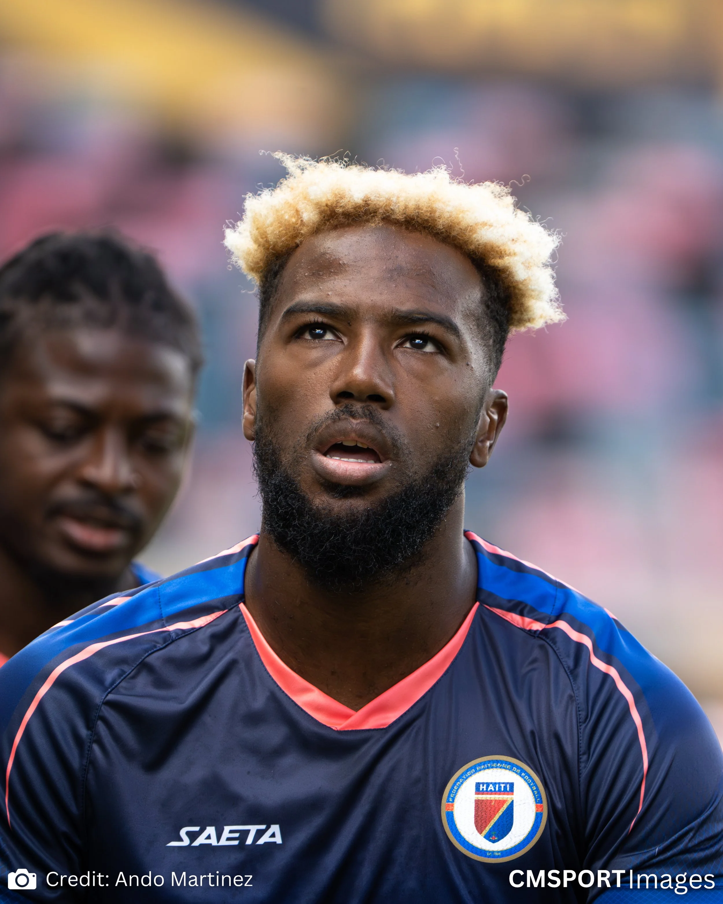 Haitian soccer player with bleached blond hair and a beard, wearing a navy blue jersey with the Haiti crest, appears exhausted. Another player is blurred in the background.