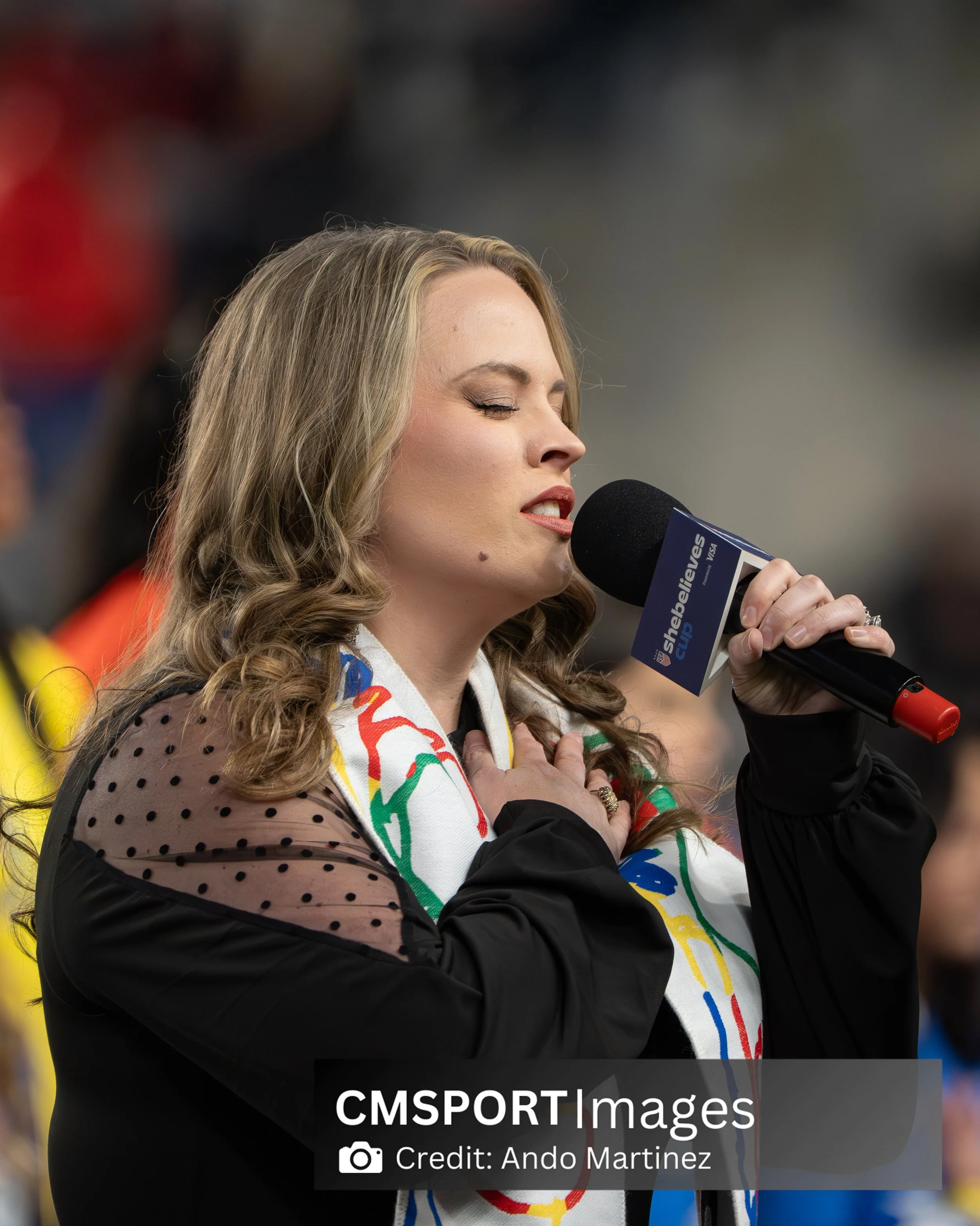 A woman with long, wavy blonde hair, eyes closed, holding her right hand on her chest, and speaking into a microphone during a sports event.