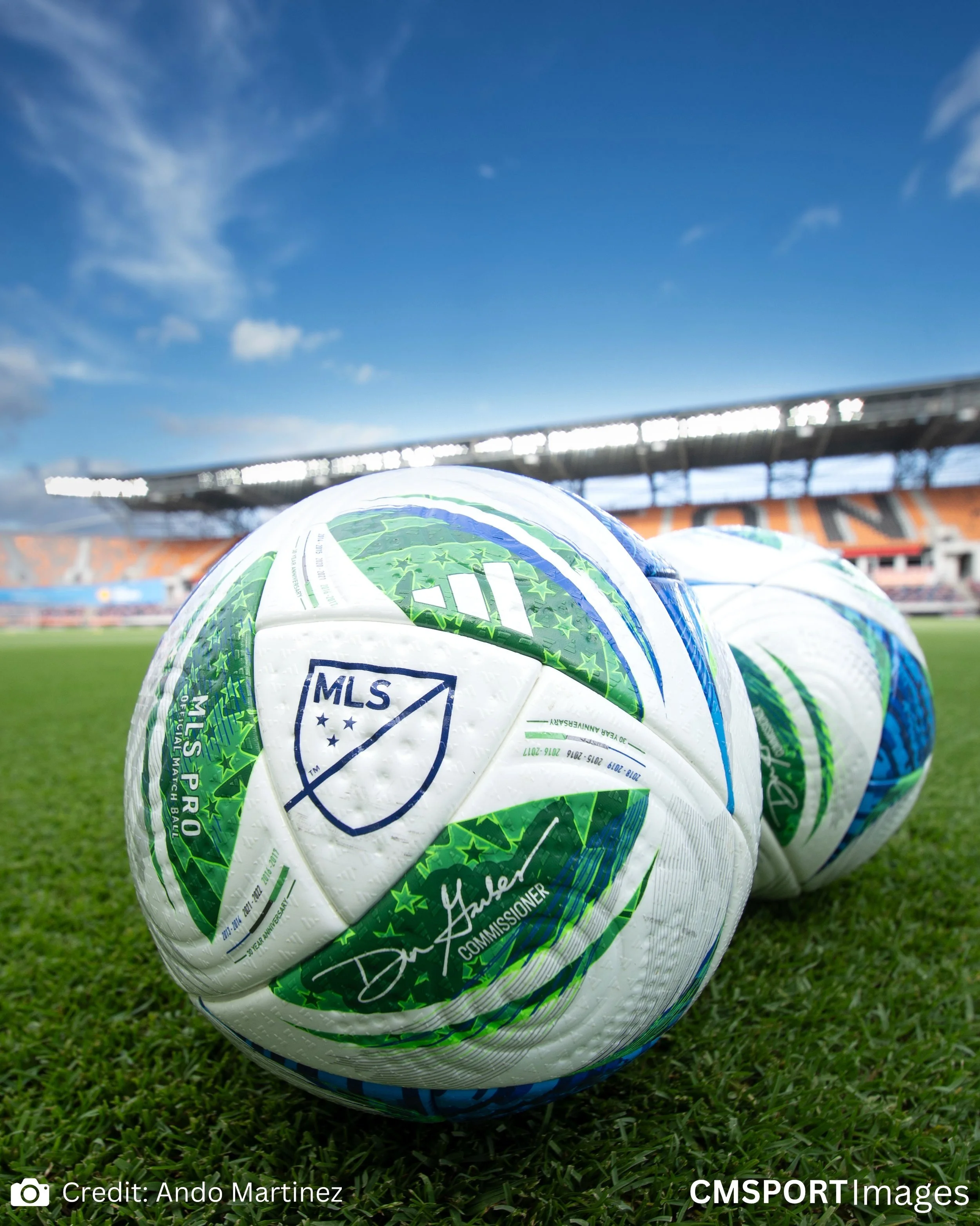 Close-up of MLS official soccer ball on green field with stadium in background under blue sky.