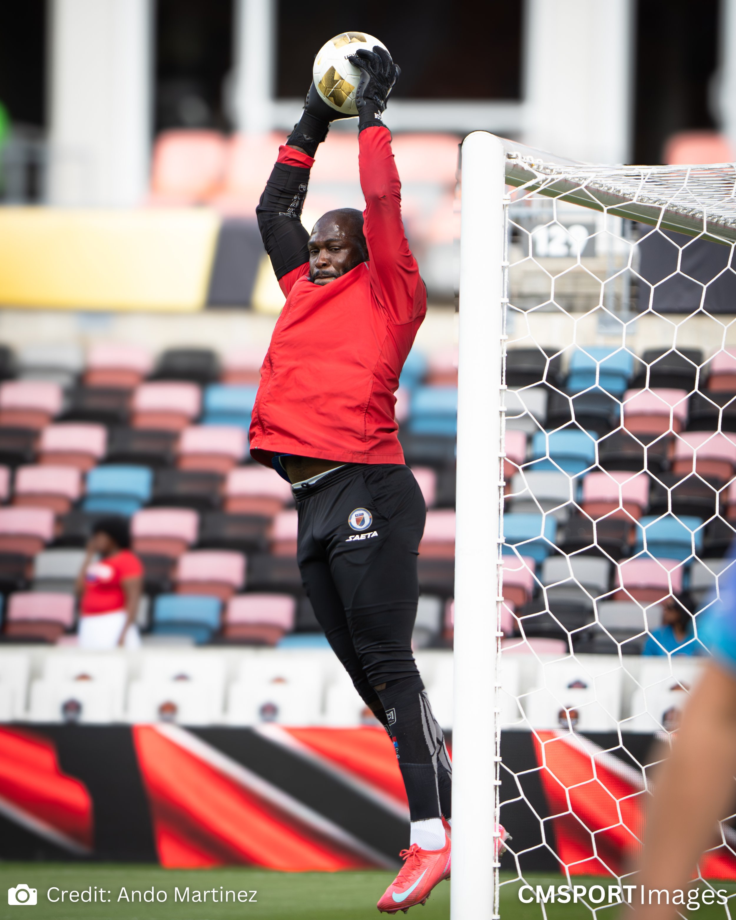 Soccer goalkeeper jumping to catch a ball during training, wearing red jersey and black pants.