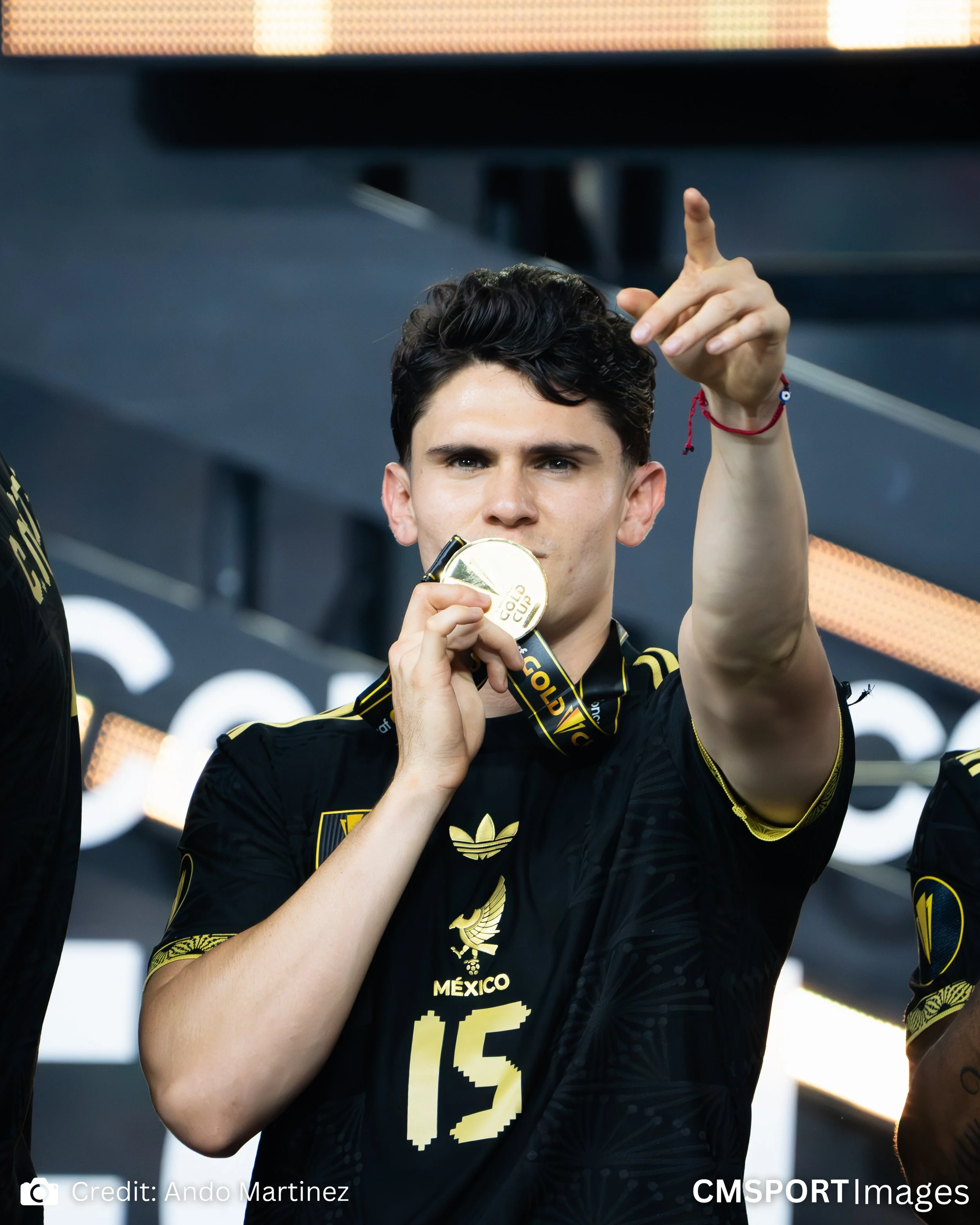 Young male athlete with dark hair holding a gold medal and making a victory gesture, wearing a black and gold sports jersey with 'Mexico' and the number 15, celebrating a win.