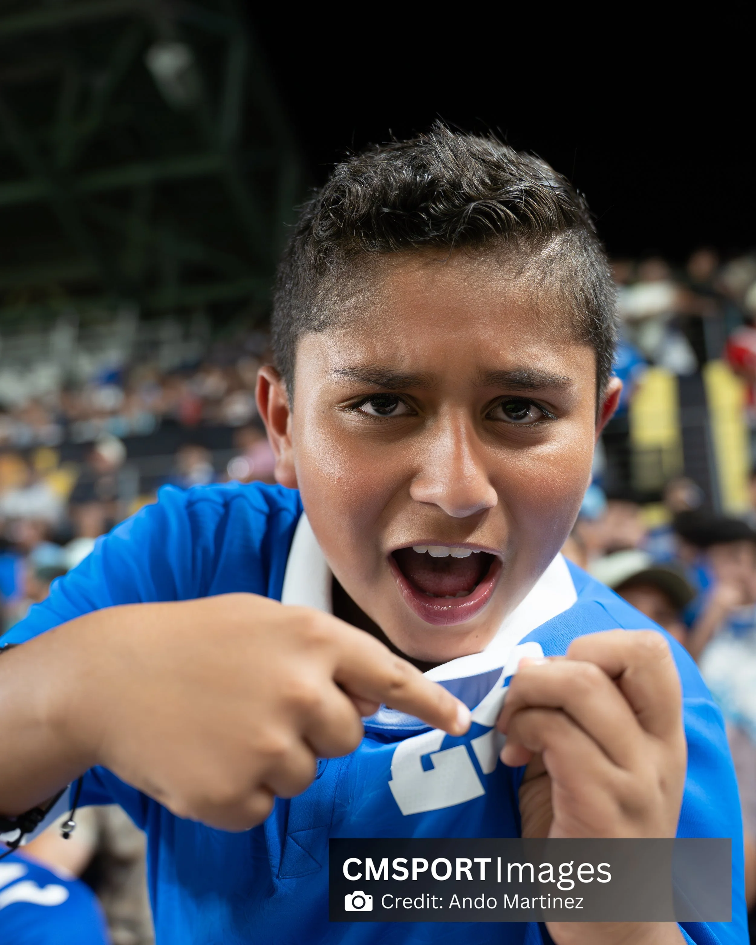A young person with short dark hair in a blue sports jersey, excitedly pointing at a jersey or shirt with a bold number on it, at a crowded sports stadium.