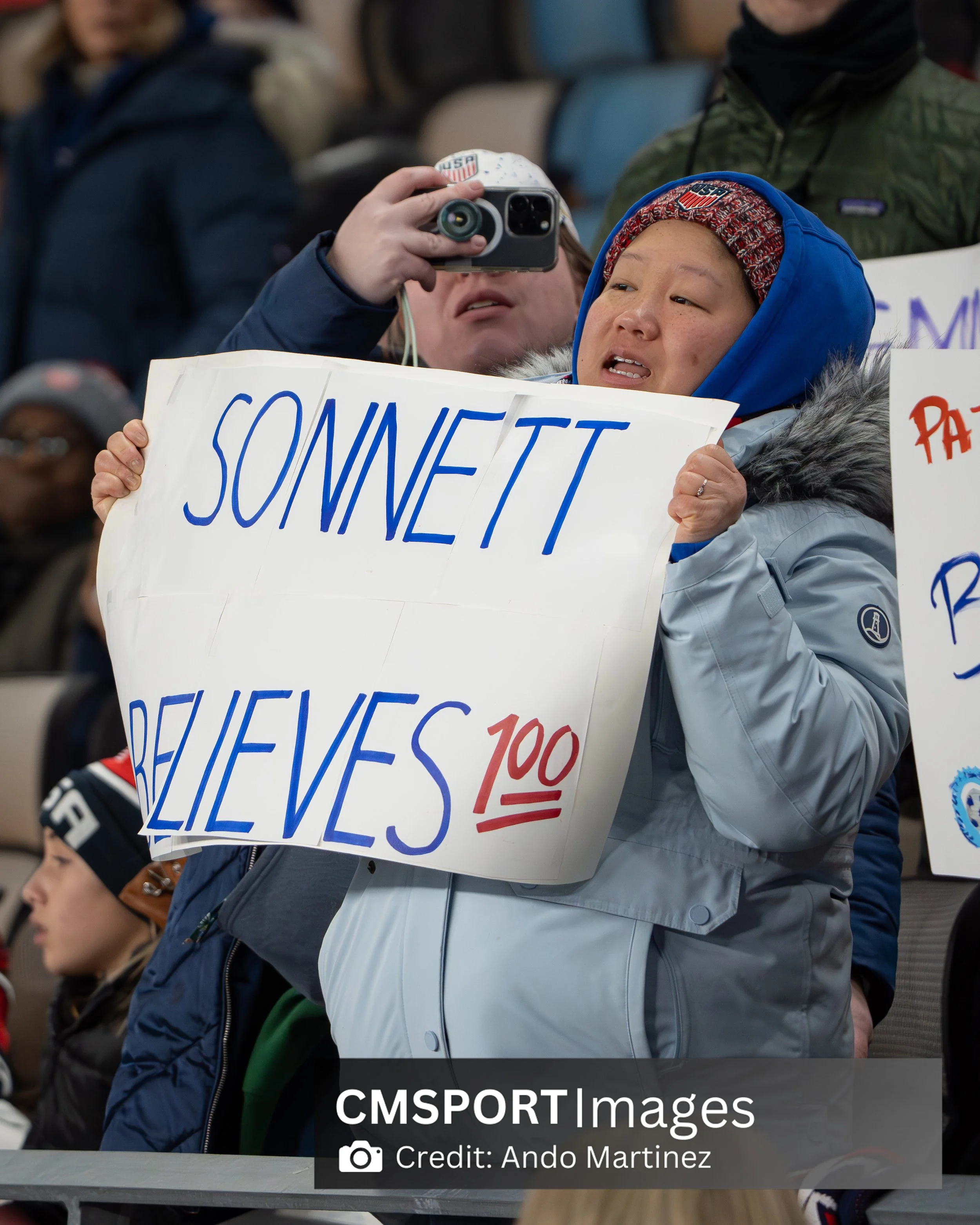 A person holding a white sign that reads 'Sonnett Believes 100%' at a crowded event, with some people taking photos and others holding signs in the background.