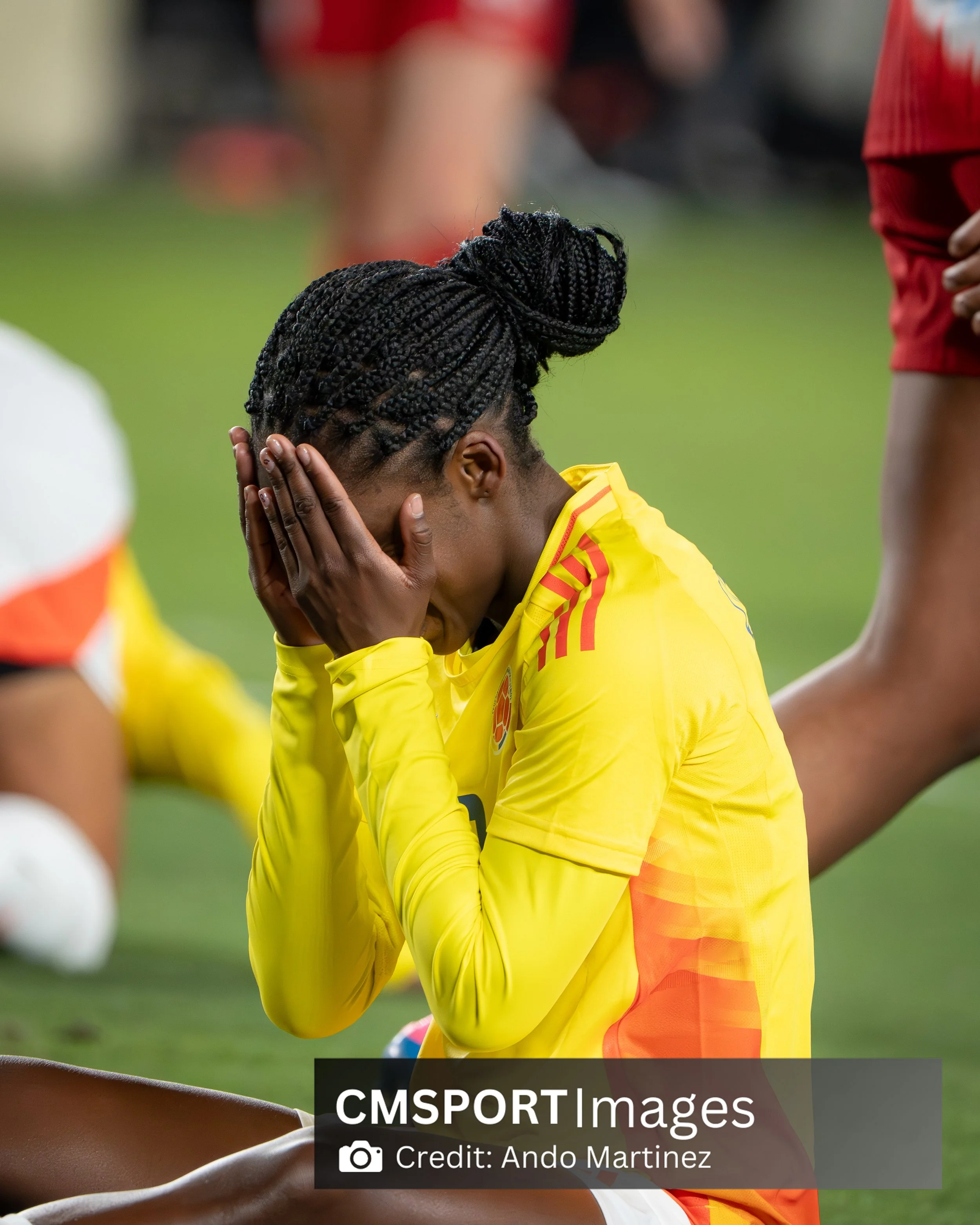 A female soccer player wearing a yellow jersey with red details, sitting on the field with her hands covering her face, appearing distressed or upset.
