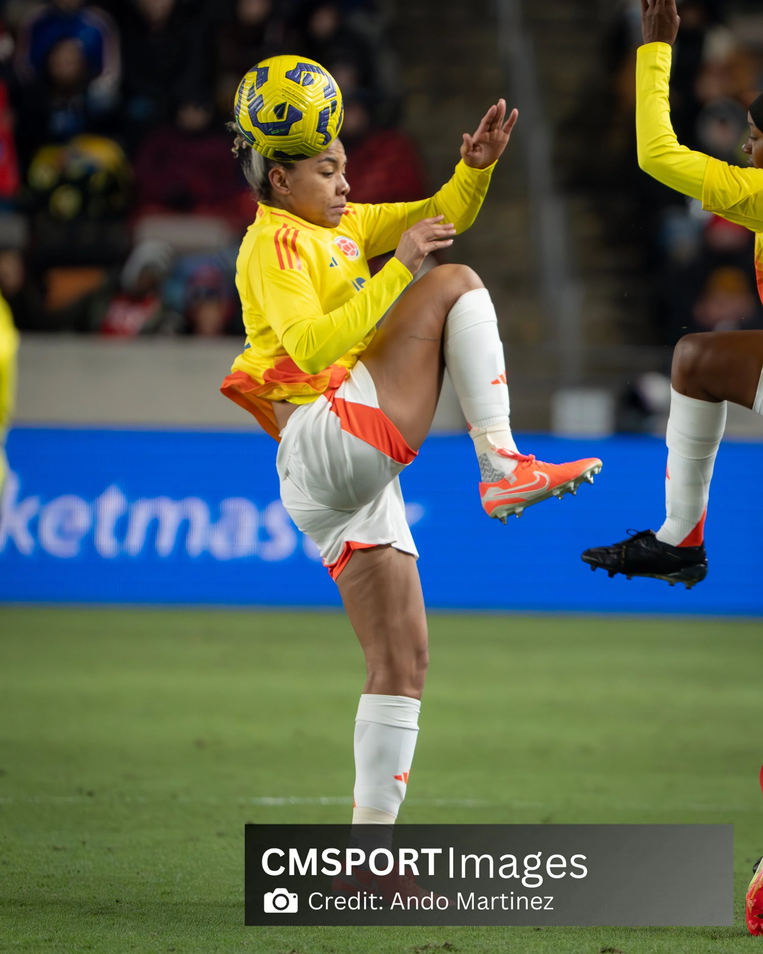 A female soccer player in a yellow jersey and white shorts balancing a yellow and purple soccer ball on her head during a match.