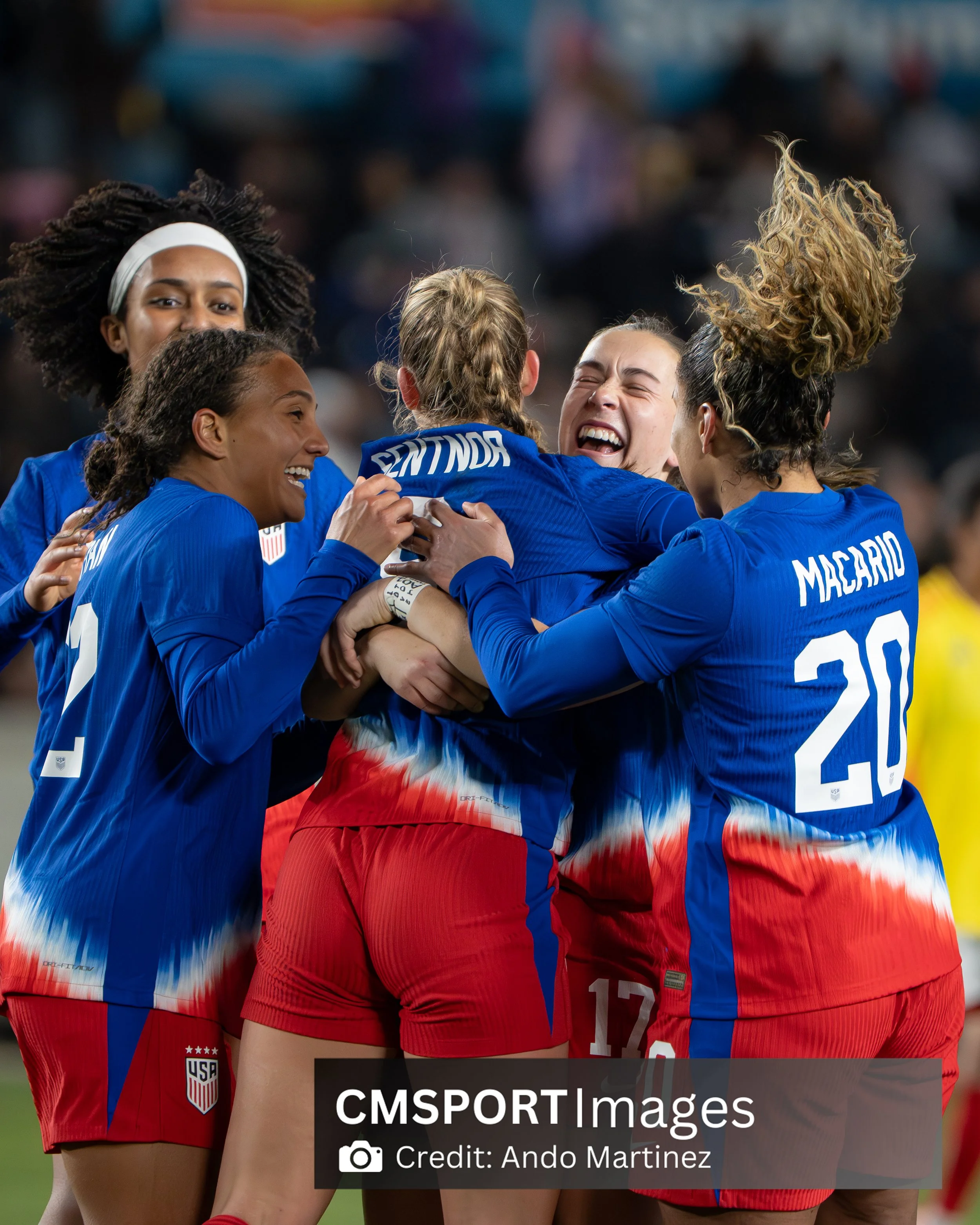 US women's national soccer team celebrating during a match, wearing blue and red uniforms.