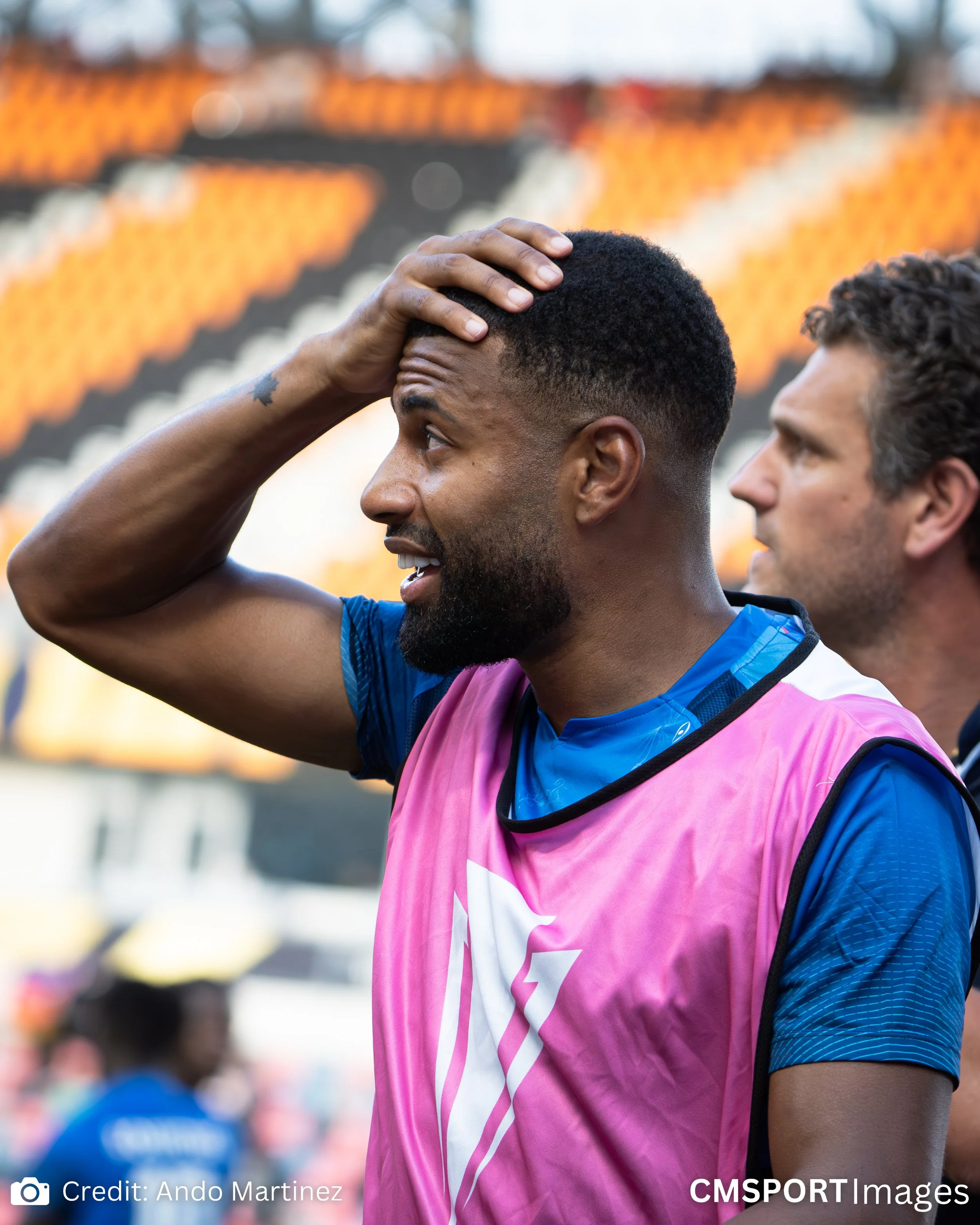 A soccer player with a beard and short hair, wearing a blue shirt and pink vest, holding his head in surprise or frustration at a stadium.