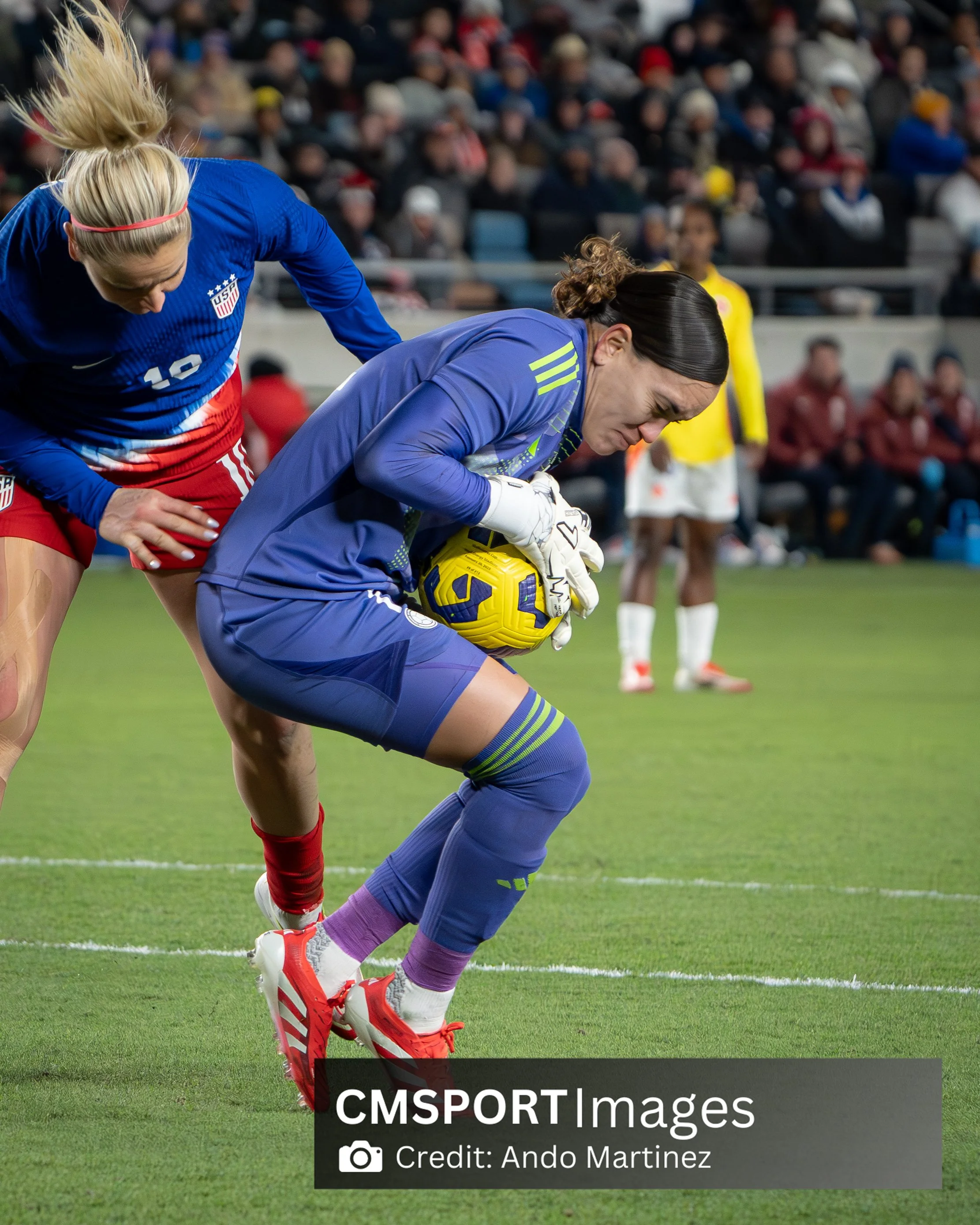 A female soccer goalkeeper in blue uniform catching a yellow soccer ball on a soccer field during a match, with a female player in a blue and red uniform attempting to challenge her and spectators in the background.